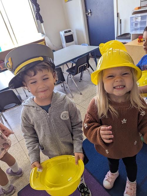 Two children wearing hats, one a captain's hat and one a hard hat, in a classroom