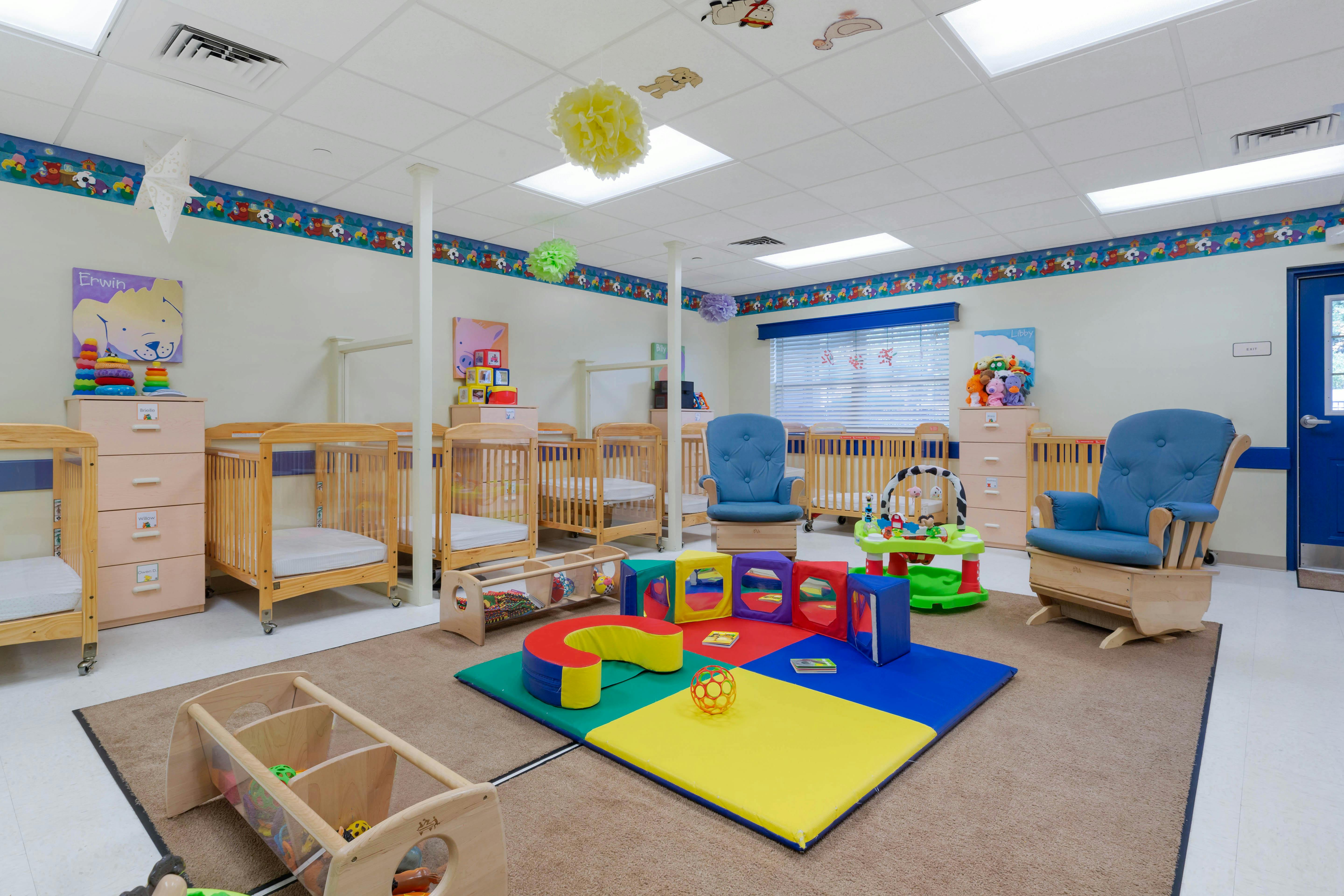 A colorful childcare room with several cribs, play mats, and two rocking chairs