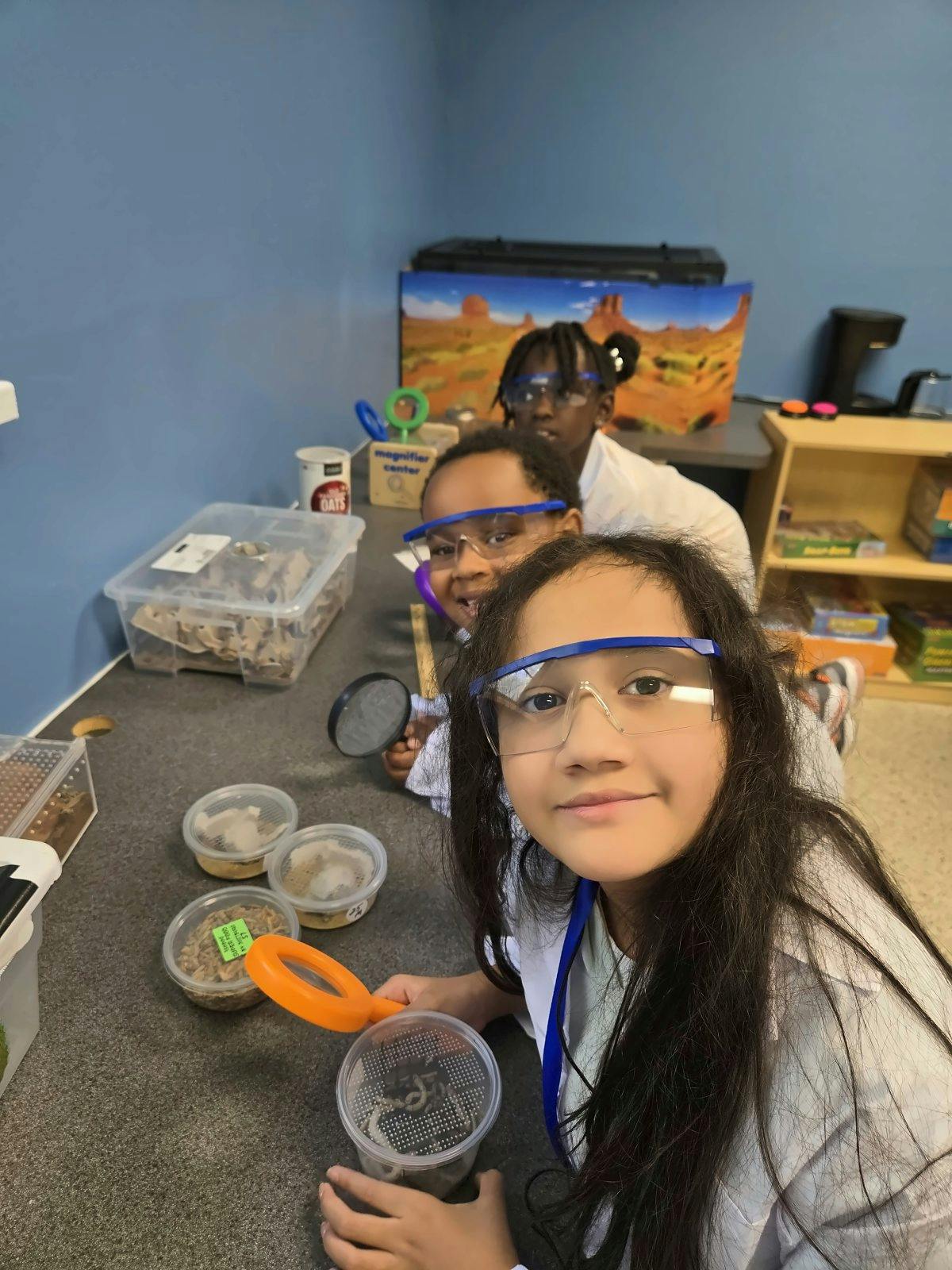 Three children wearing safety goggles examining insects in a science classroom