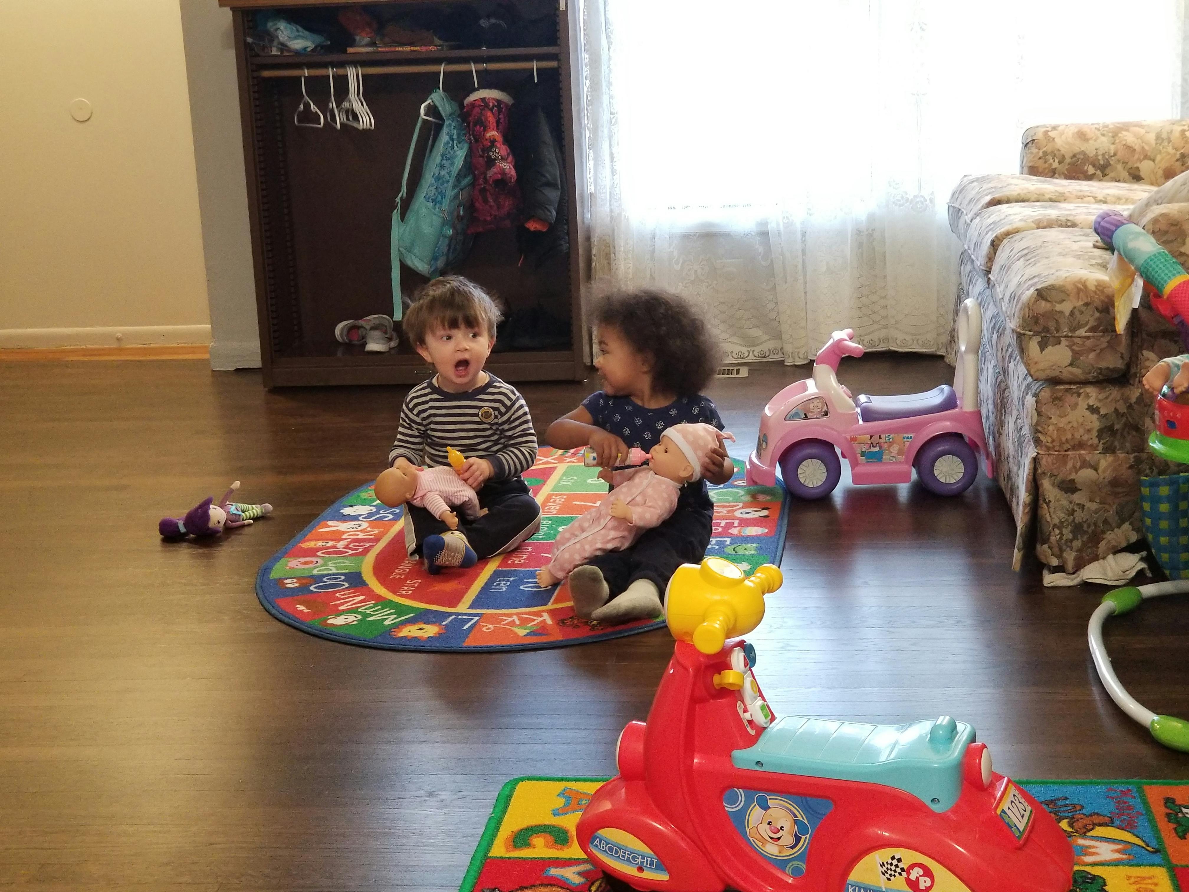 Two children sitting on a playmat holding dolls