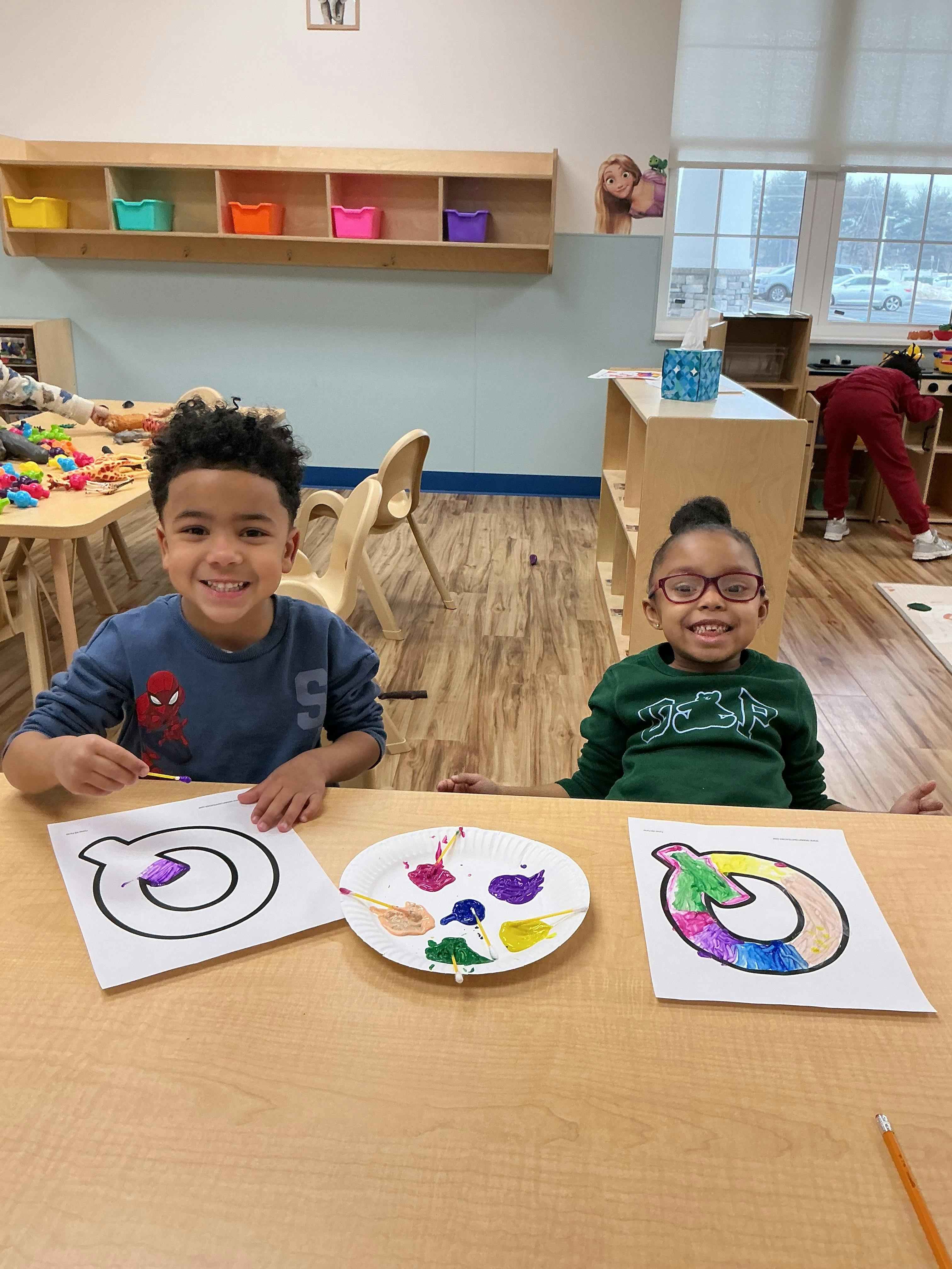 Two children at a table, smiling and painting the letter Q