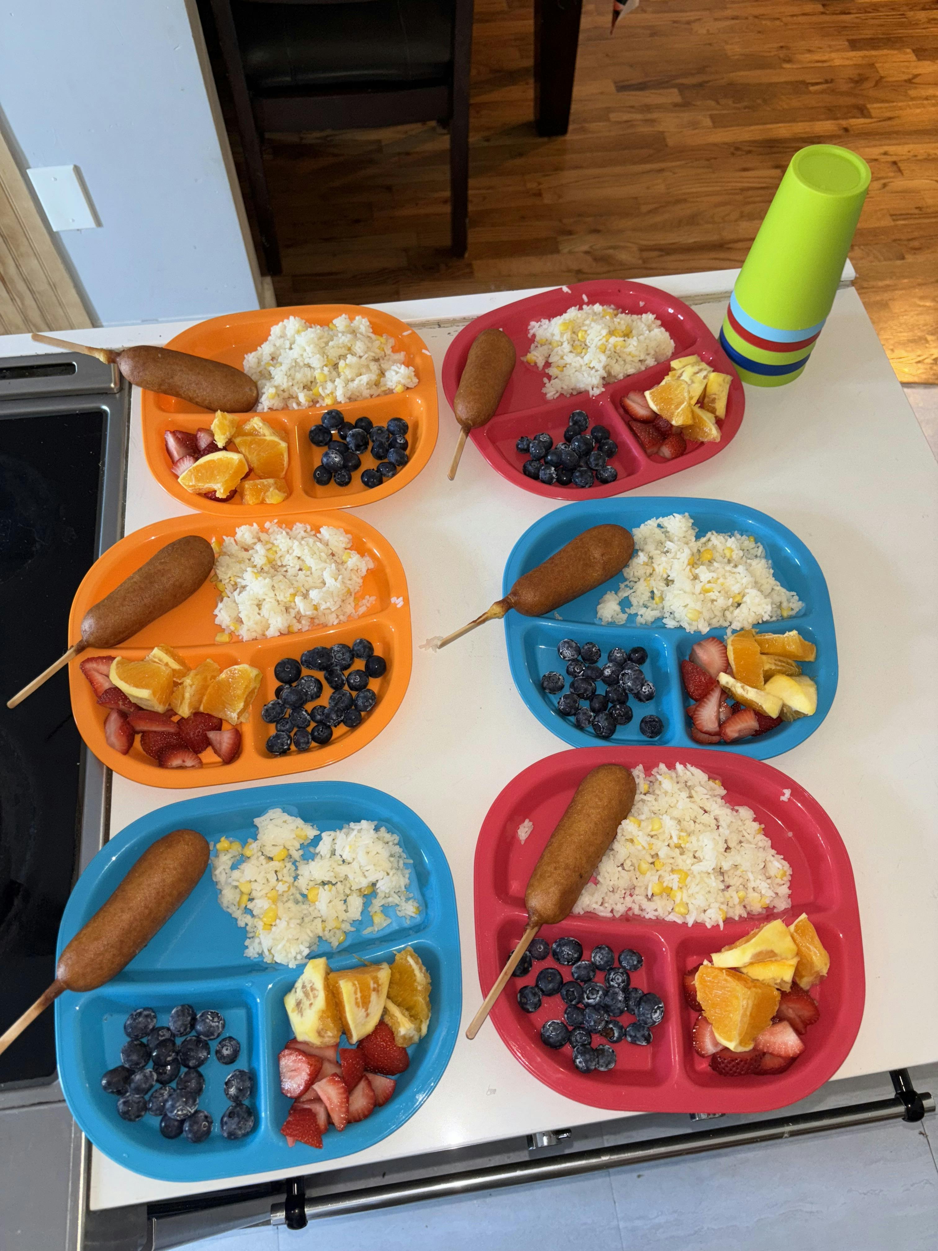 Six divided plates with corn dogs, rice, blueberries, and fruit