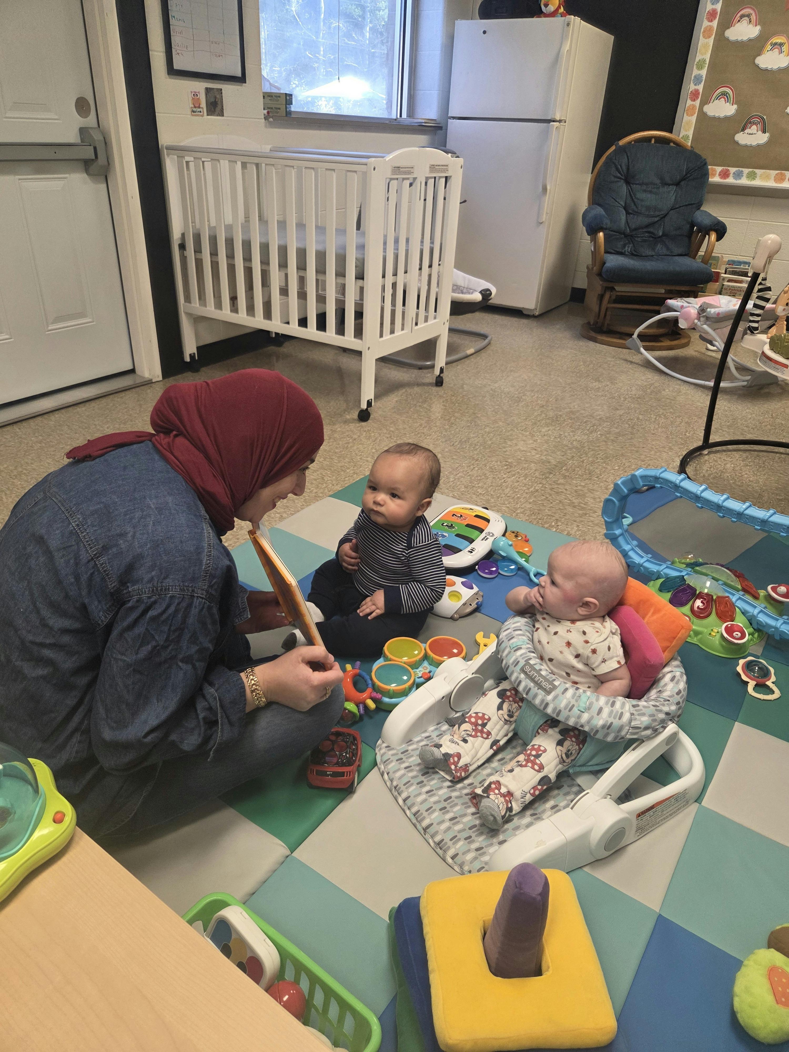 Person in a red hijab reading to two babies on a playmat in a childcare room