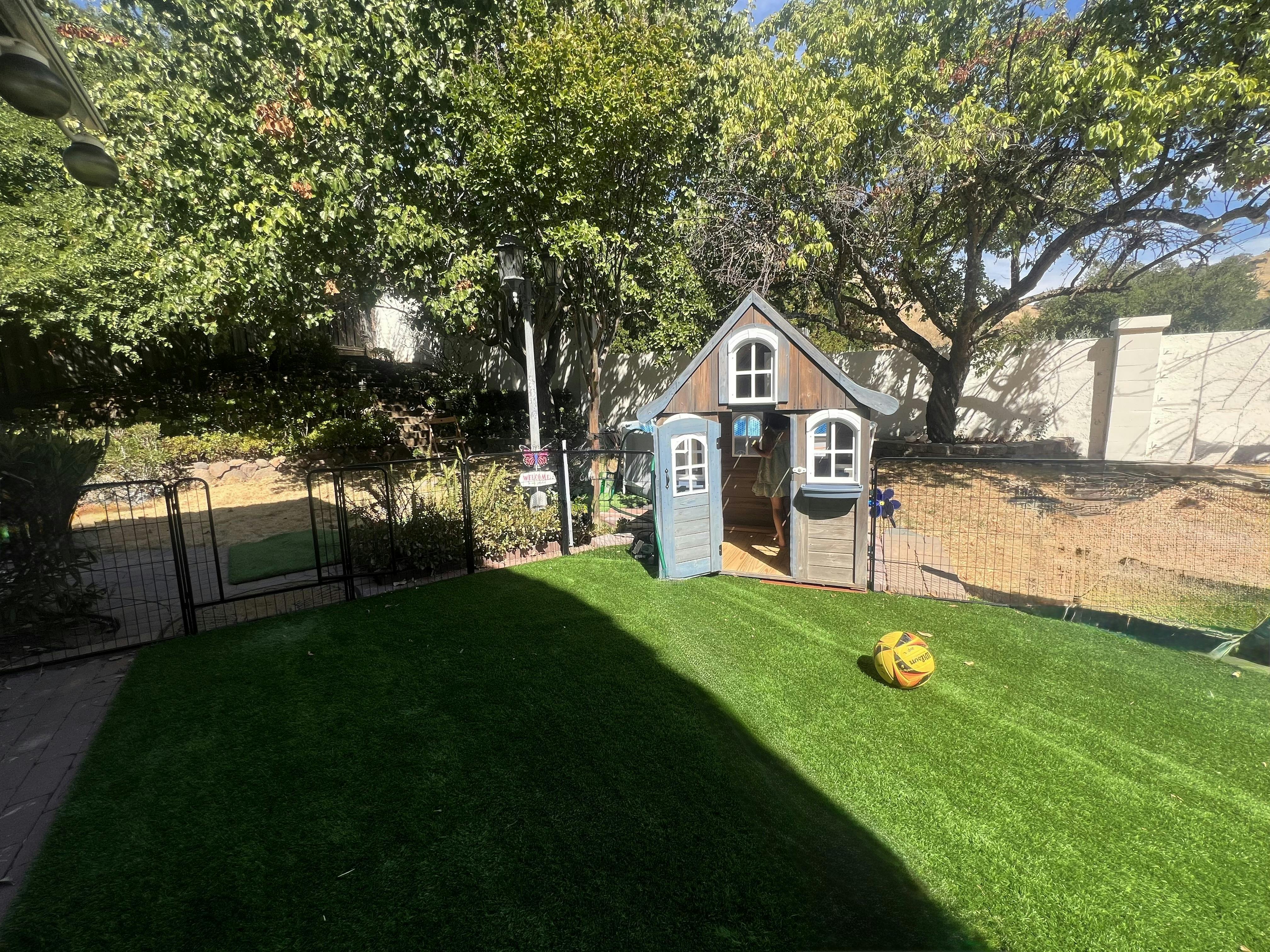 Child standing inside a playhouse on a green lawn with a yellow ball