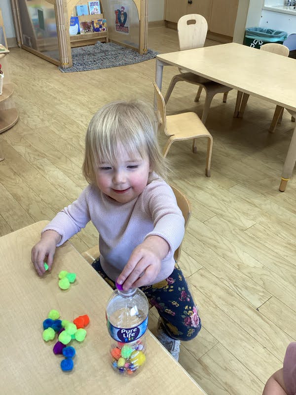 Child putting colorful pom-poms into a water bottle at a table in a classroom