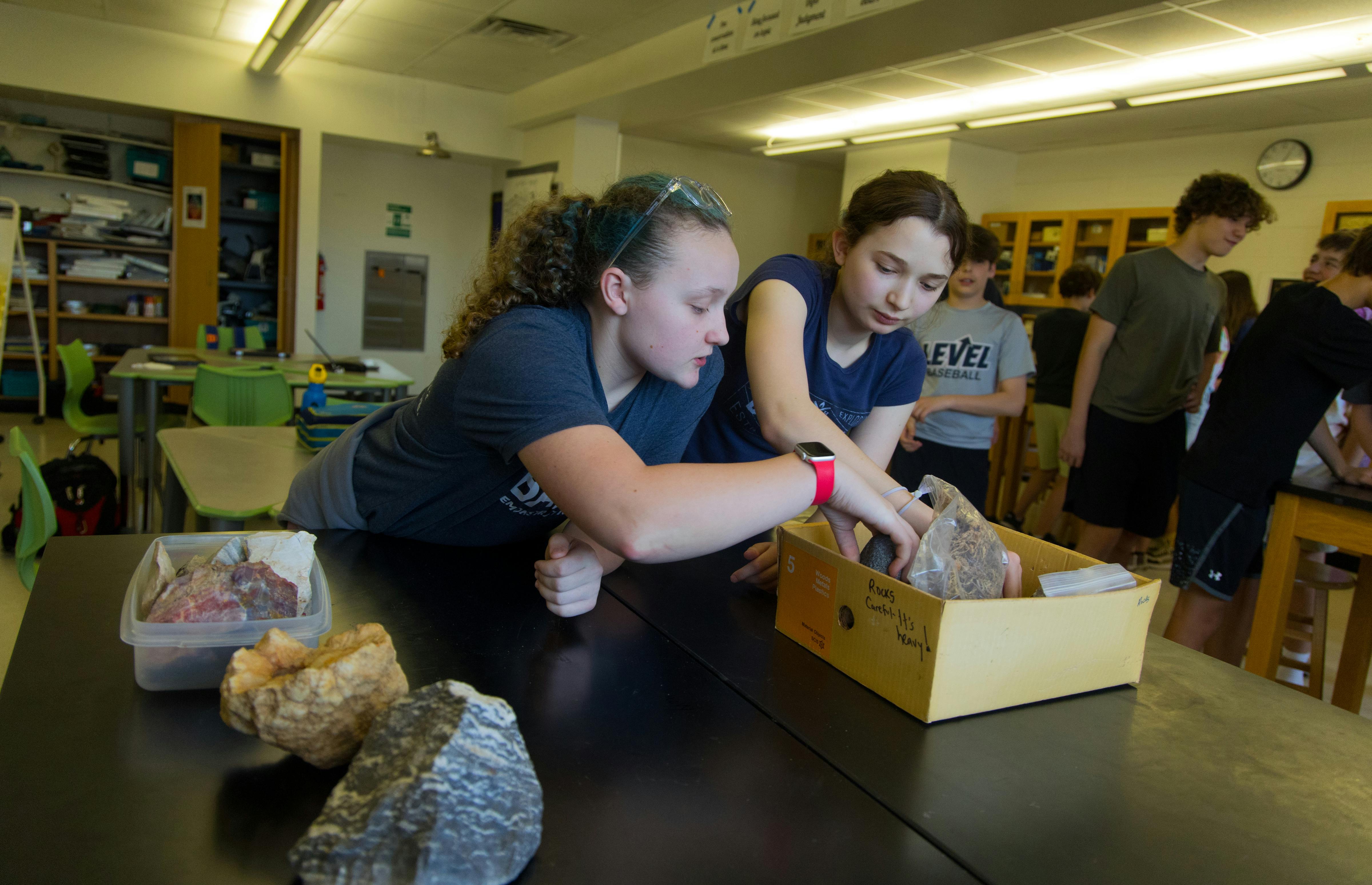 Two people examining items in a box on a table in a classroom