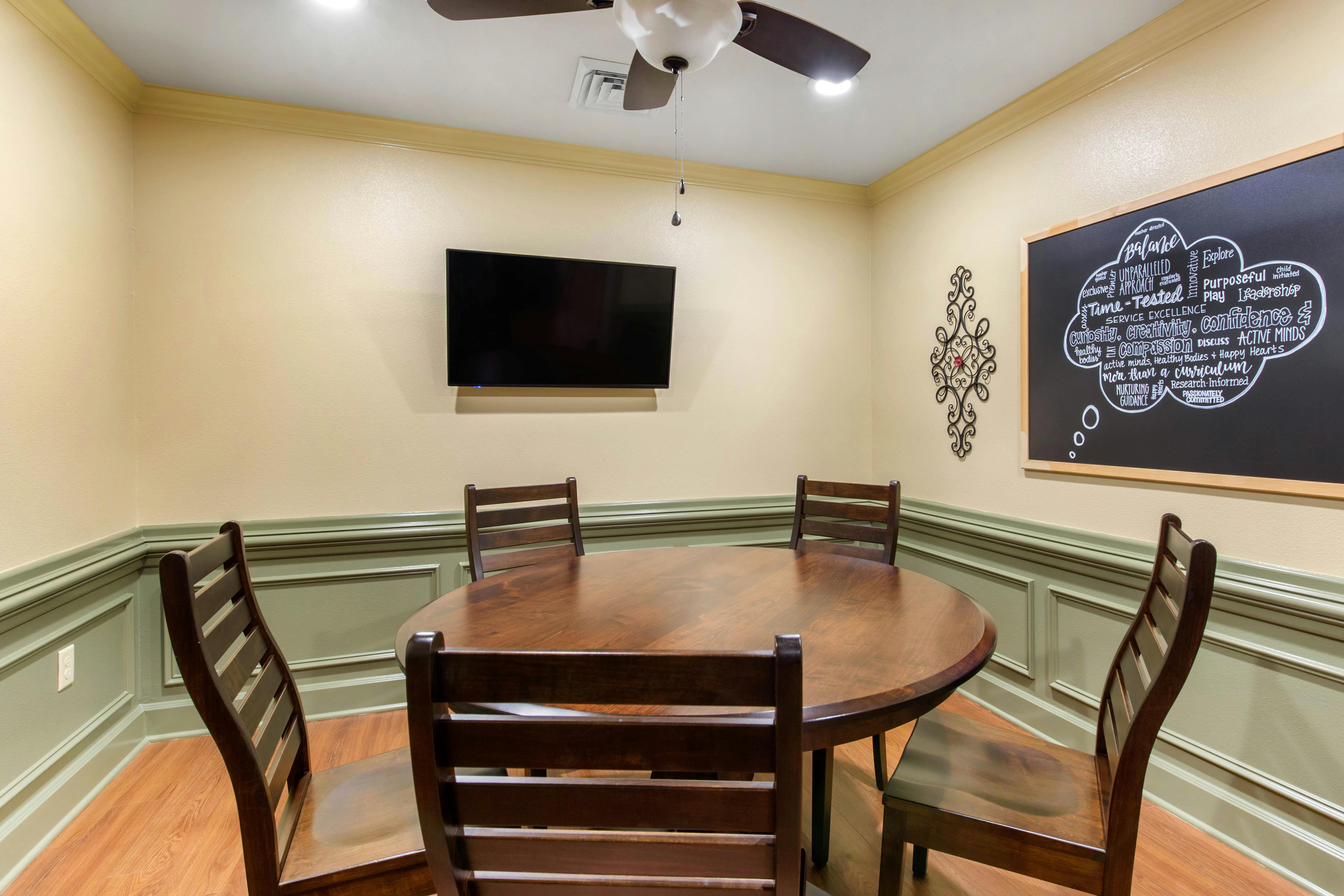 A room with a round wooden table, four chairs, a wall-mounted TV, and a chalkboard