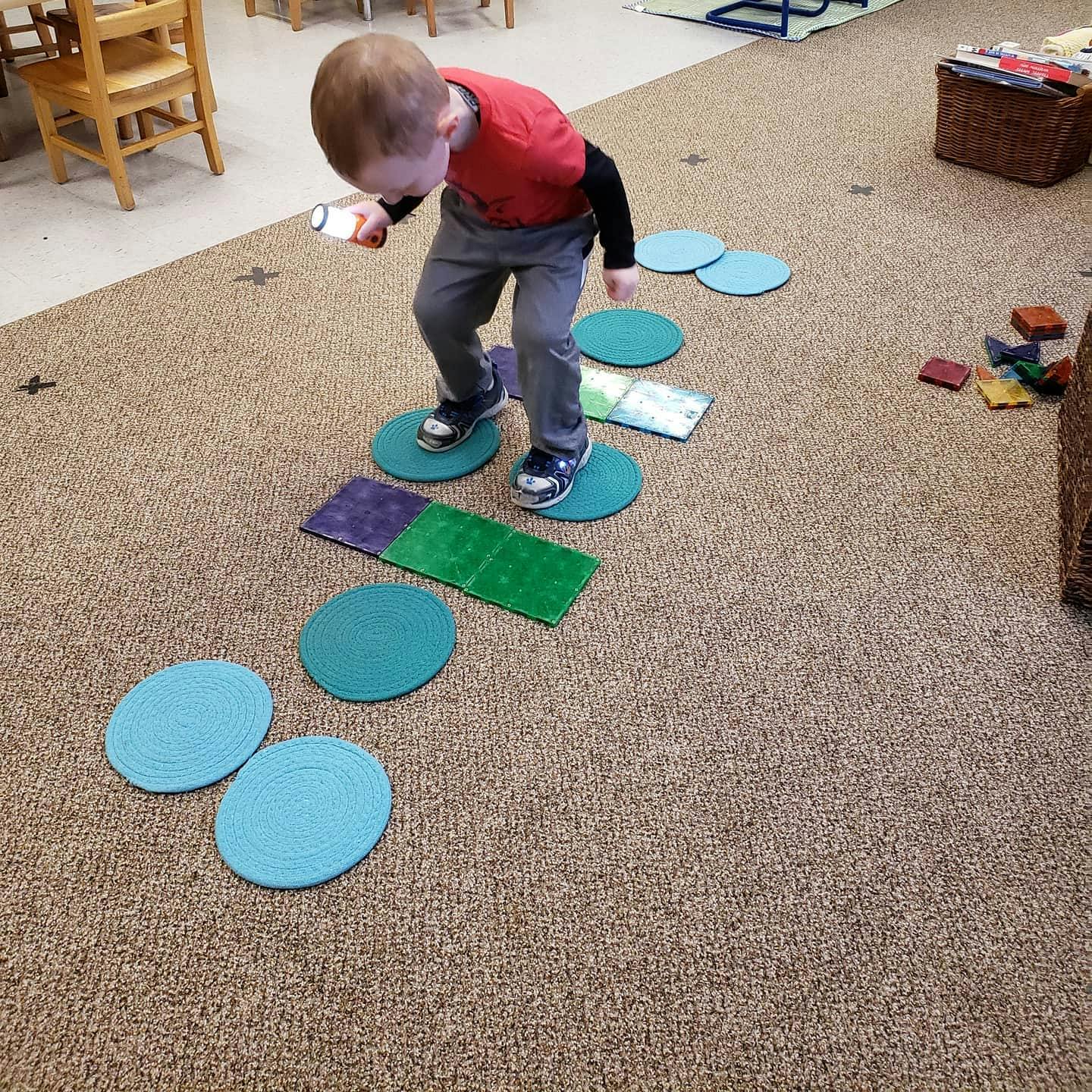 Child standing on blue circles on a carpeted floor, holding a flashlight