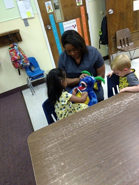 Person and two children playing with a blue monster puppet in a classroom