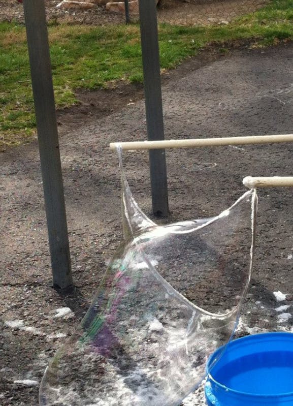 Large bubble being formed by two sticks and a string over a blue bucket