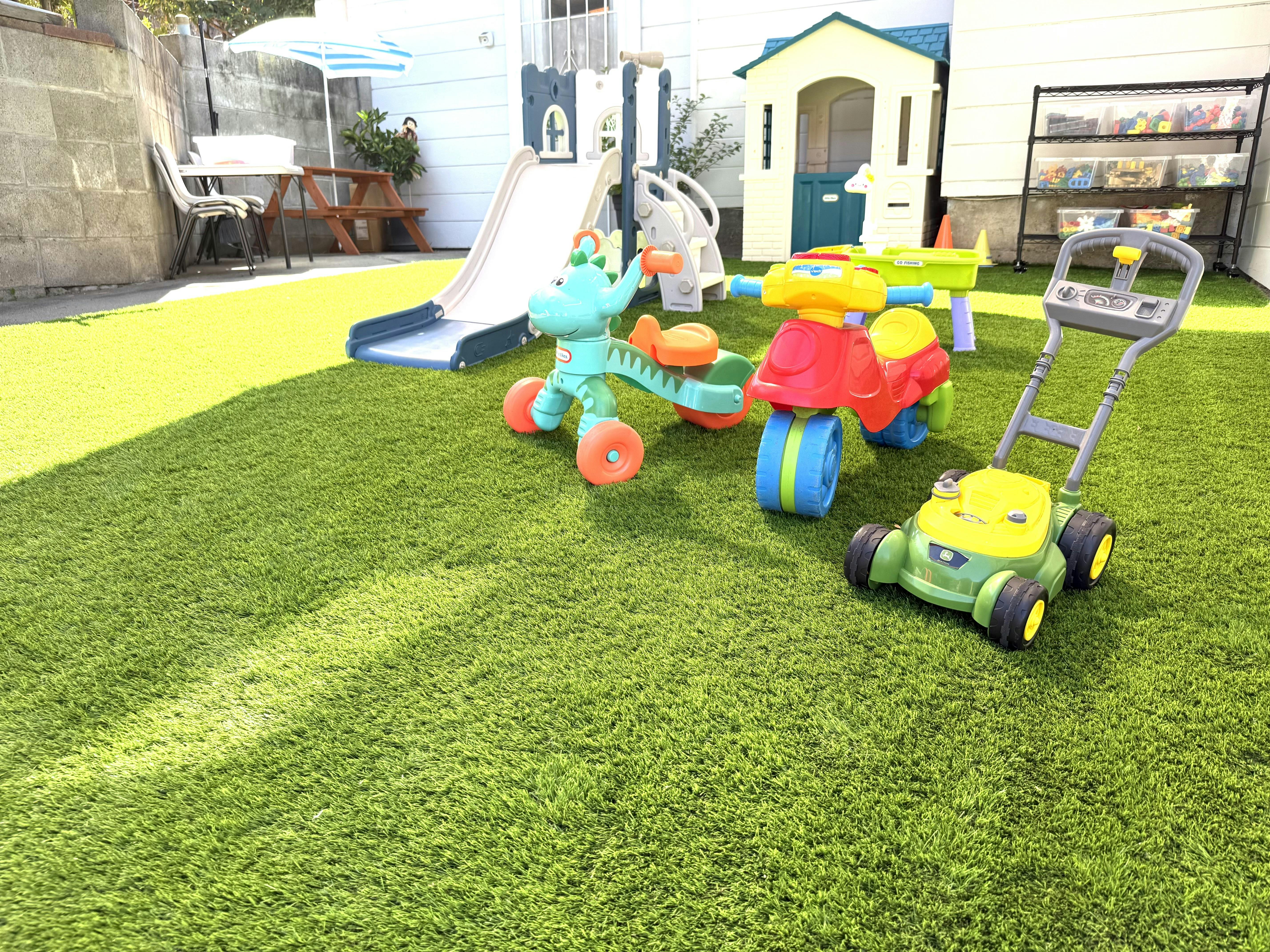 Plastic ride-on toys and a toy lawn mower on artificial grass in a backyard