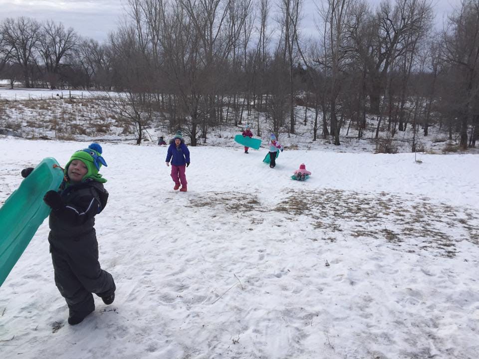 Child carrying a sled up a snowy hill with other children sledding in background