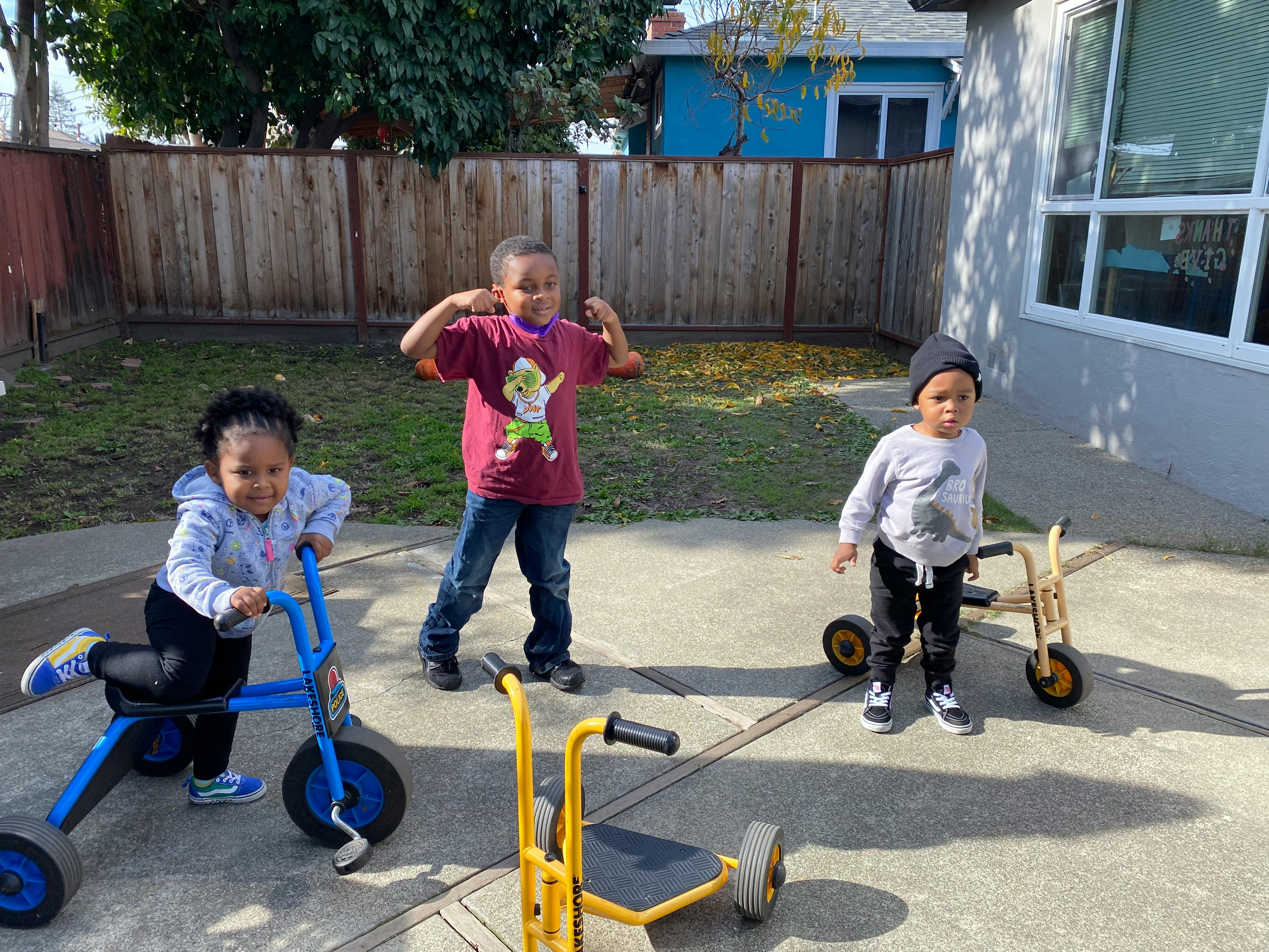 Three children with two tricycles and one scooter on a paved area