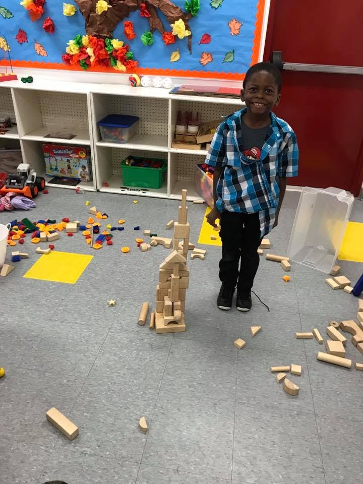 Child standing next to a tall wooden block structure in a classroom