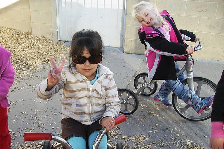 Child in sunglasses on tricycle making peace sign, another child on bicycle