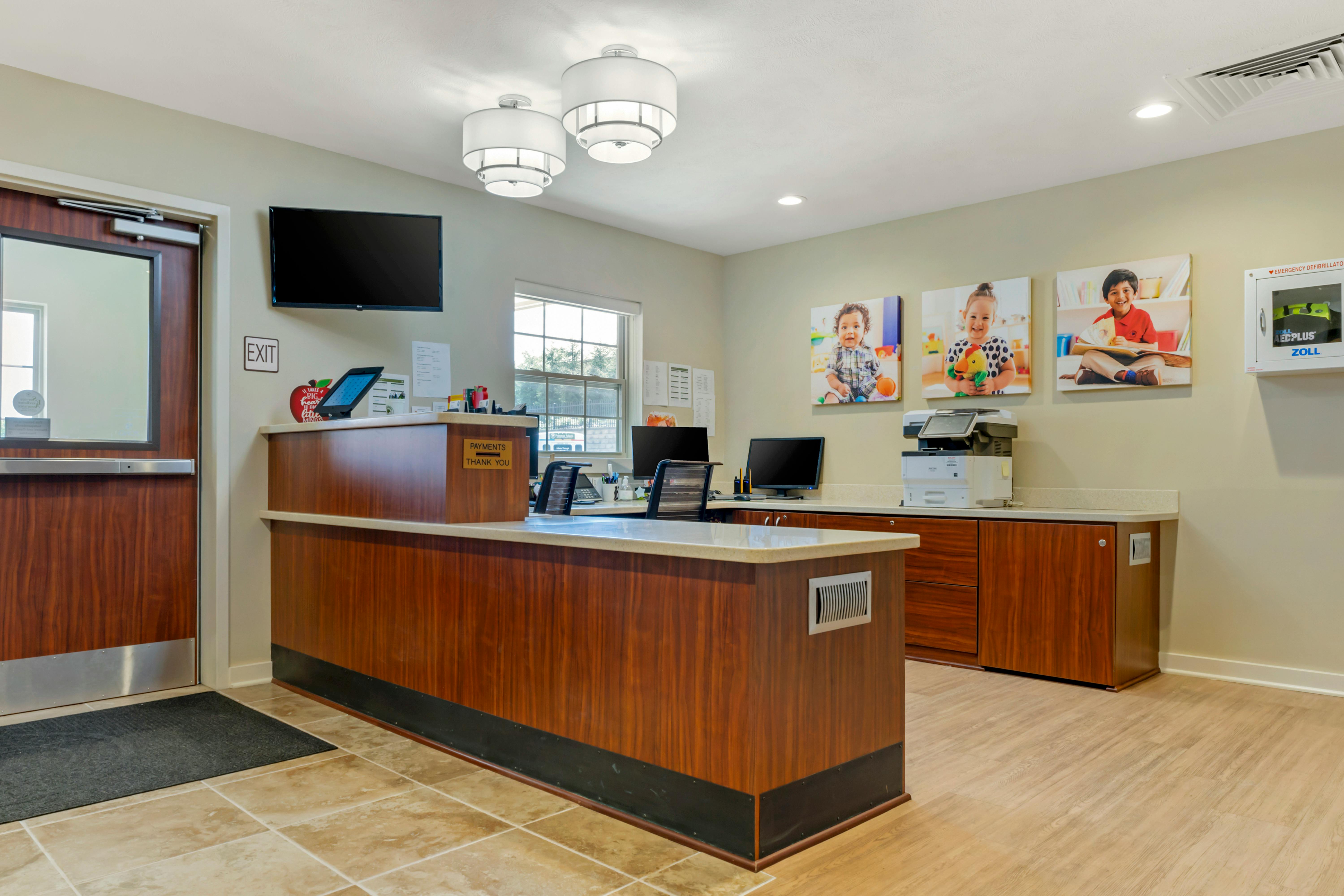 Reception area with a wooden desk, office chairs, two computers, and child portraits on the wall