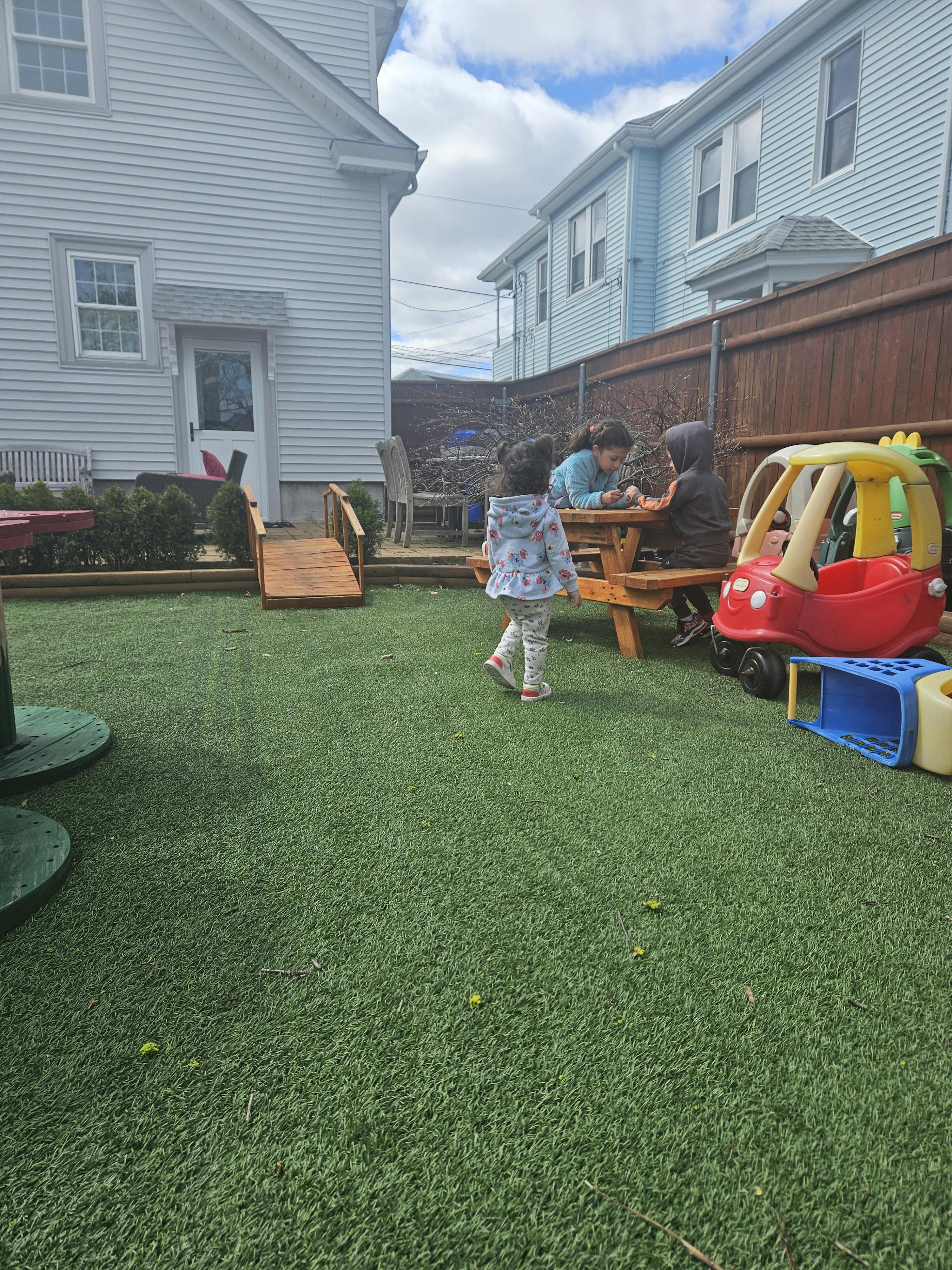 Child walking on artificial grass while other children sit at a picnic table