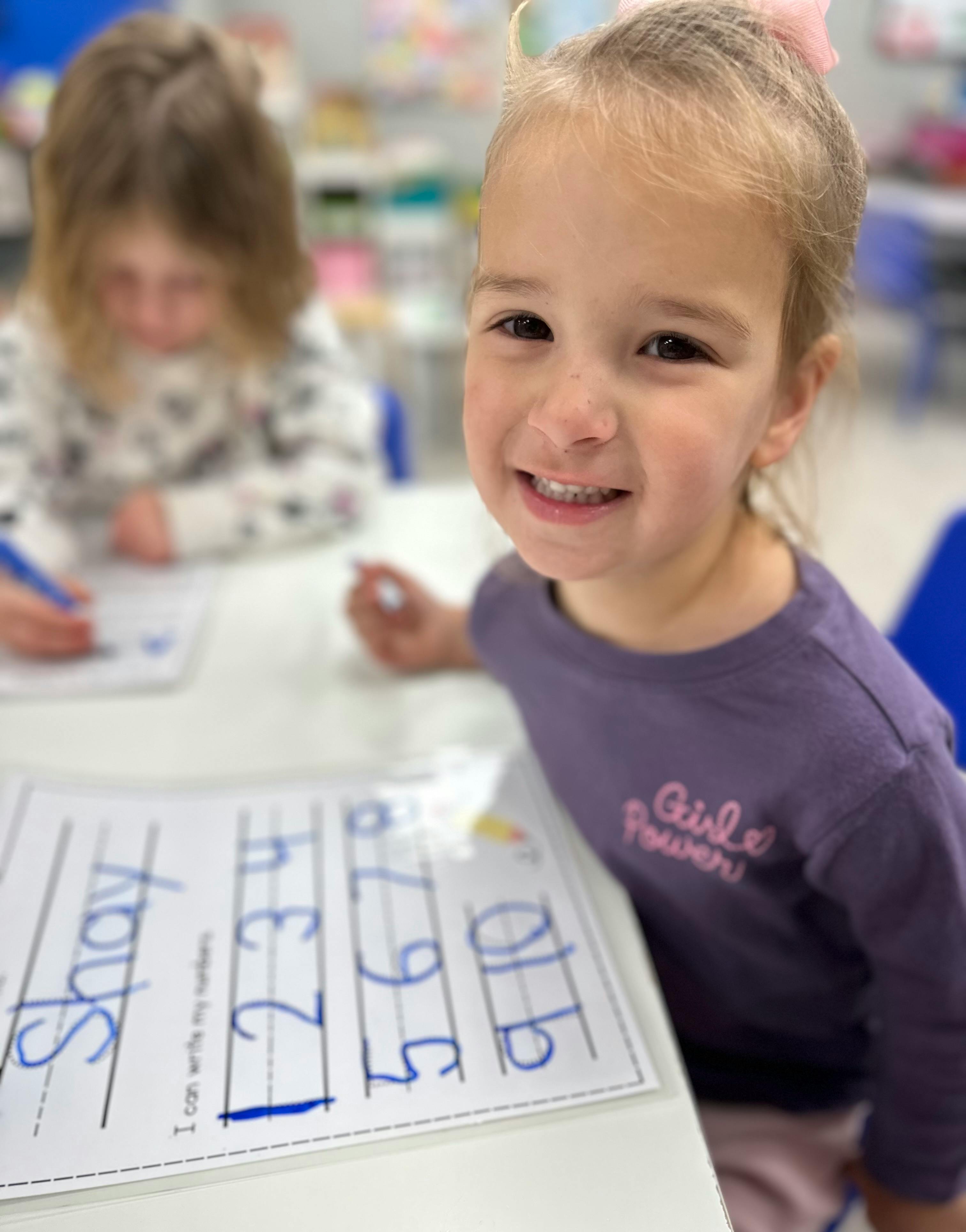 Child smiling at desk with another child writing in background