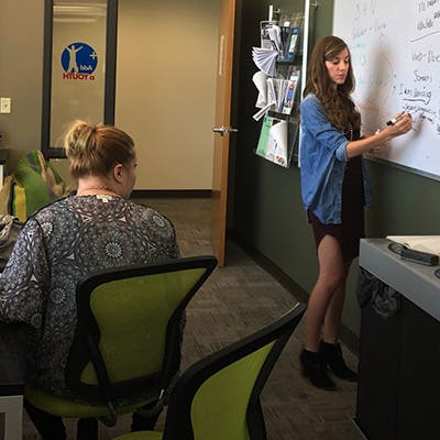 Person writing on a whiteboard while another person sits at a desk