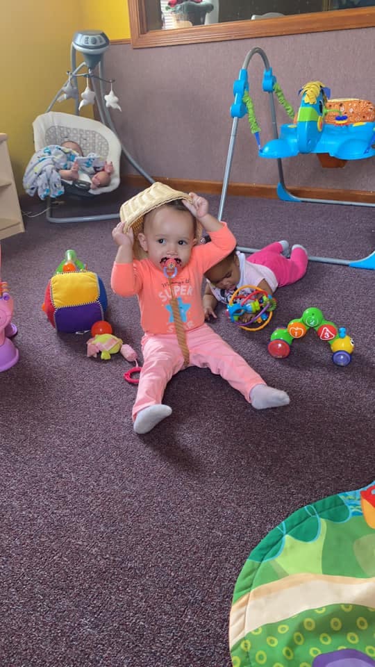 Child with pacifier holding a hat on head, another child on floor with toys