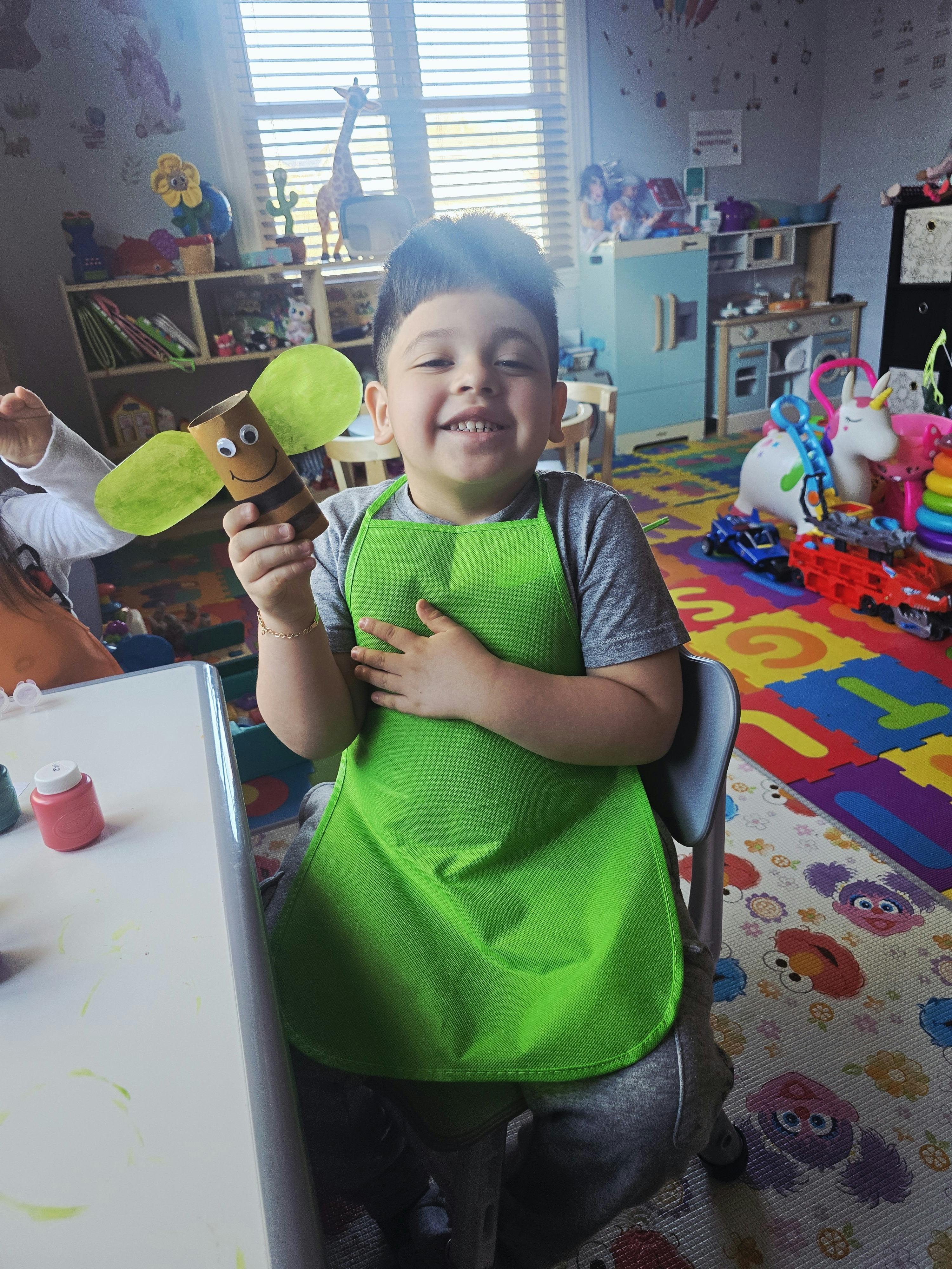Child in a green apron holding a craft bee made from a toilet paper roll