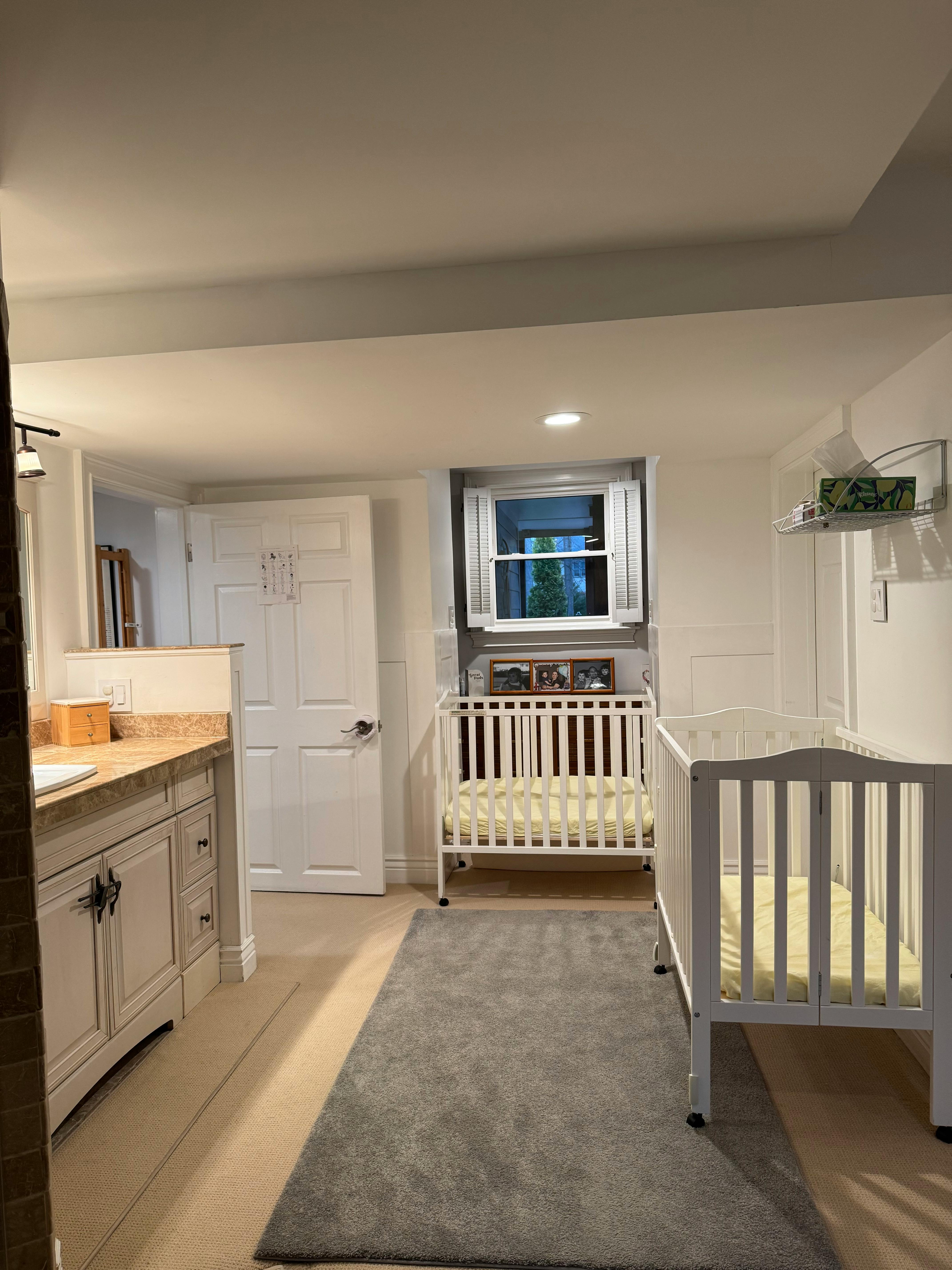 Two white cribs in a room with a window and a counter