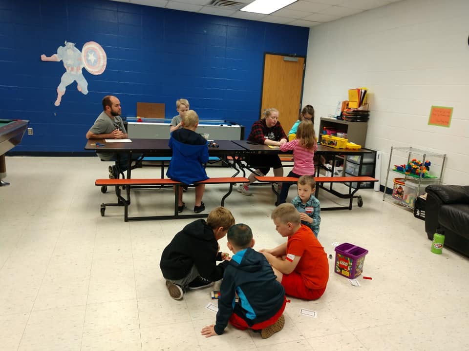 Adults and children gathered around tables and on the floor in a room