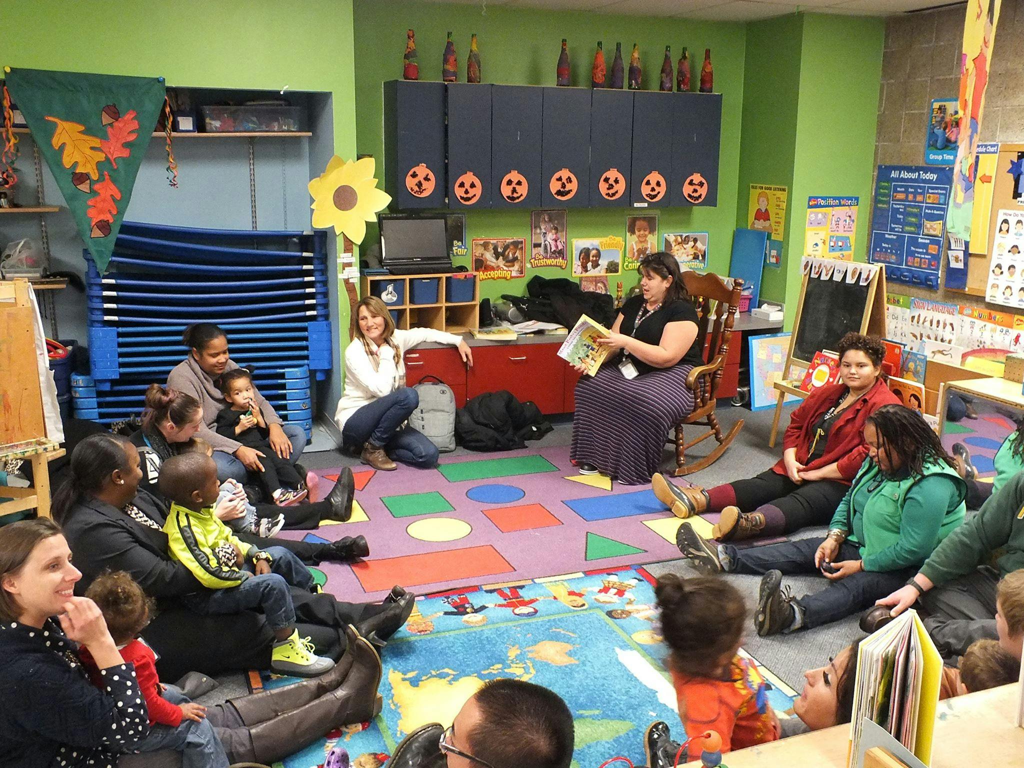 Adults and children sitting on the floor in a classroom listening to a person read