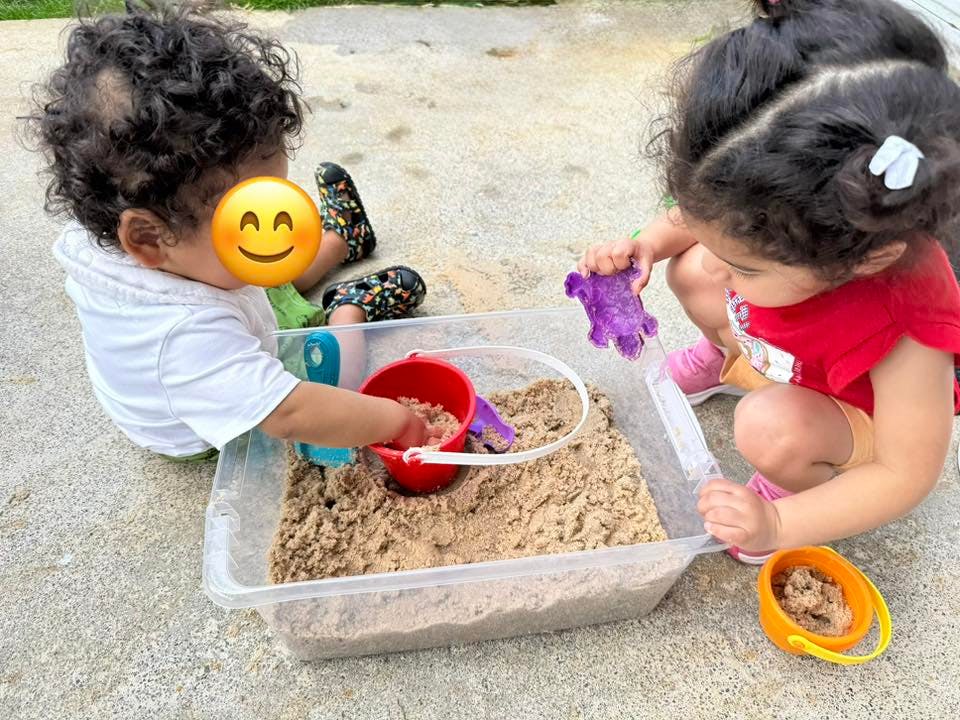 Two children playing with sand and toys in a clear plastic bin