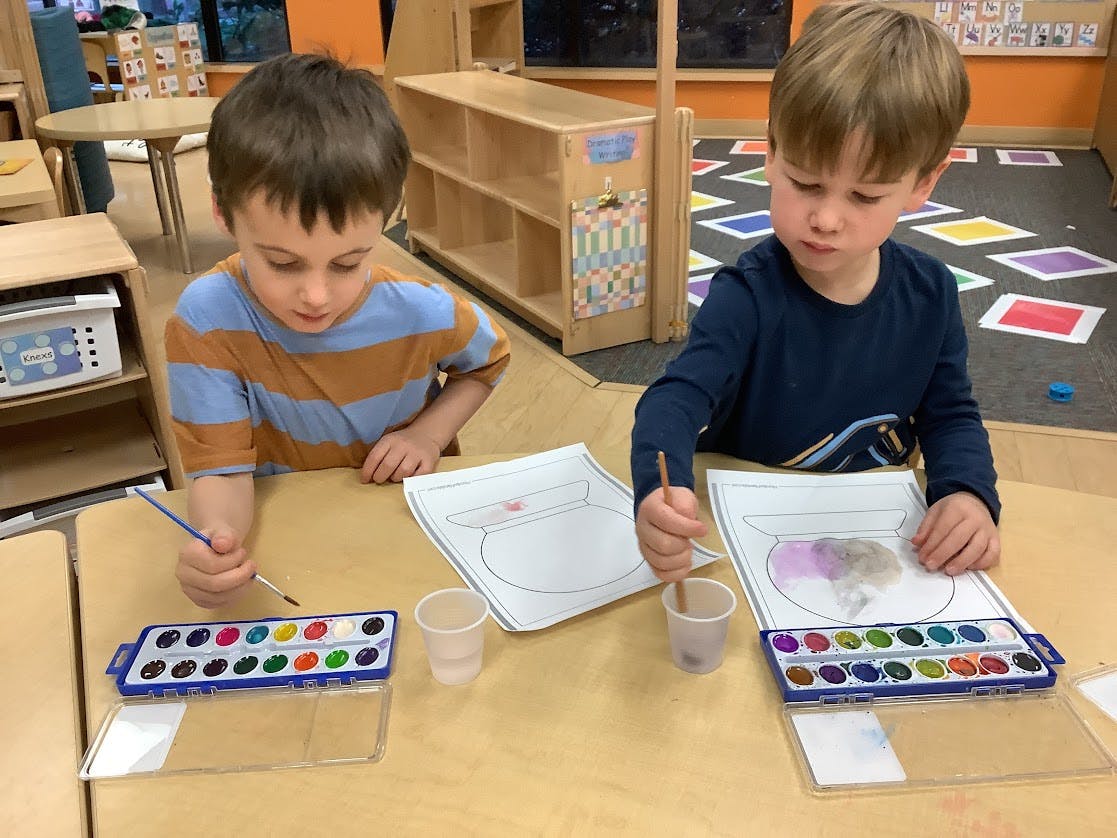 Two children painting fishbowl drawings with watercolors at a table in a classroom