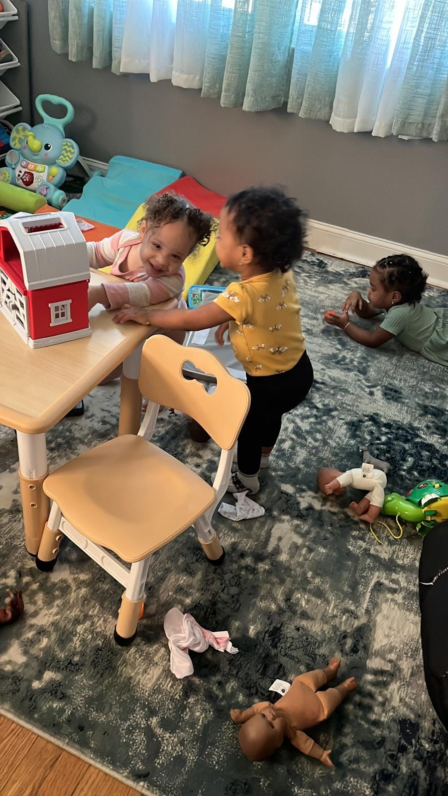Two children playing with a toy house at a small table, another child on the floor