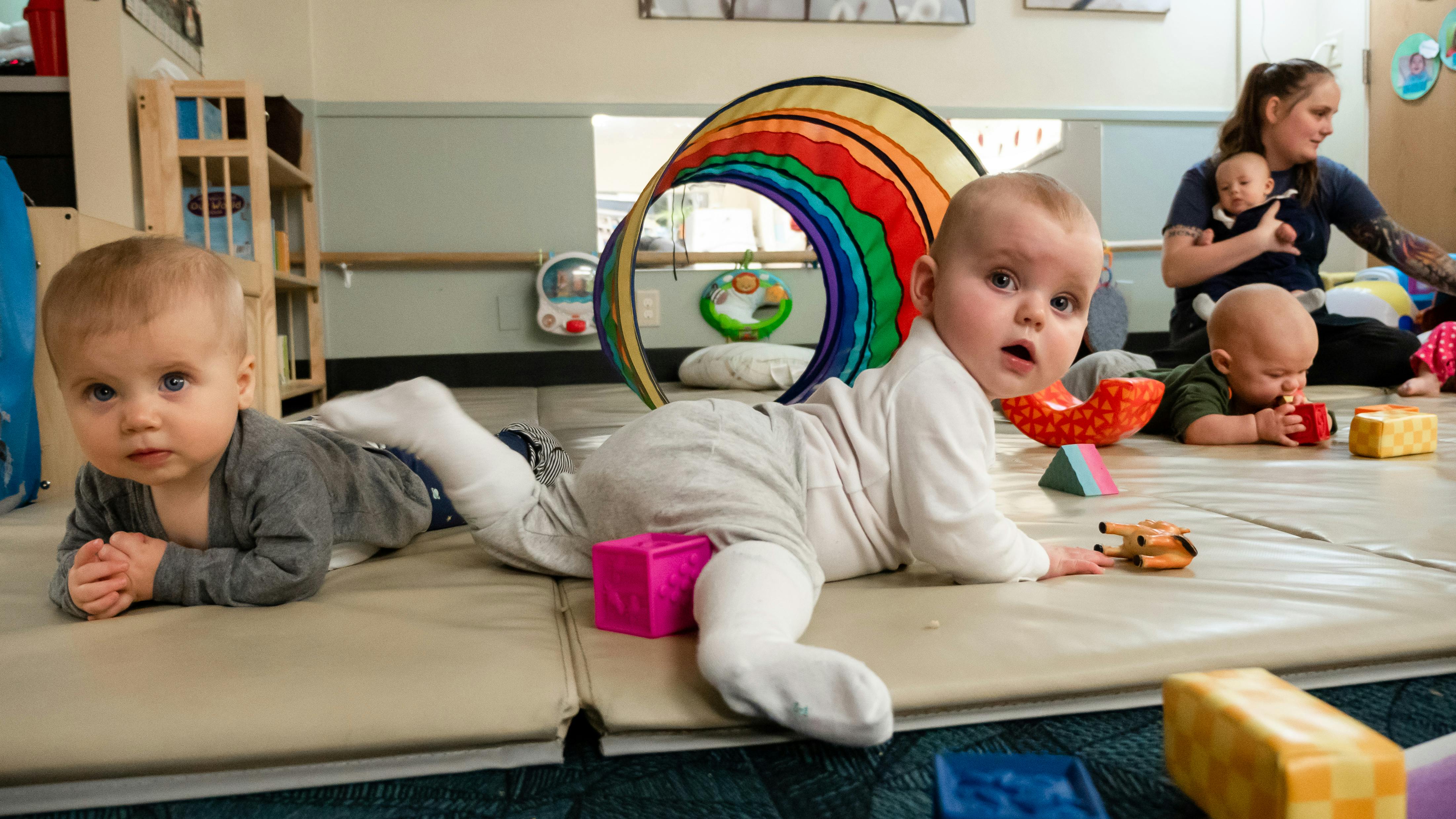Two babies on play mats with a person holding a baby in the background