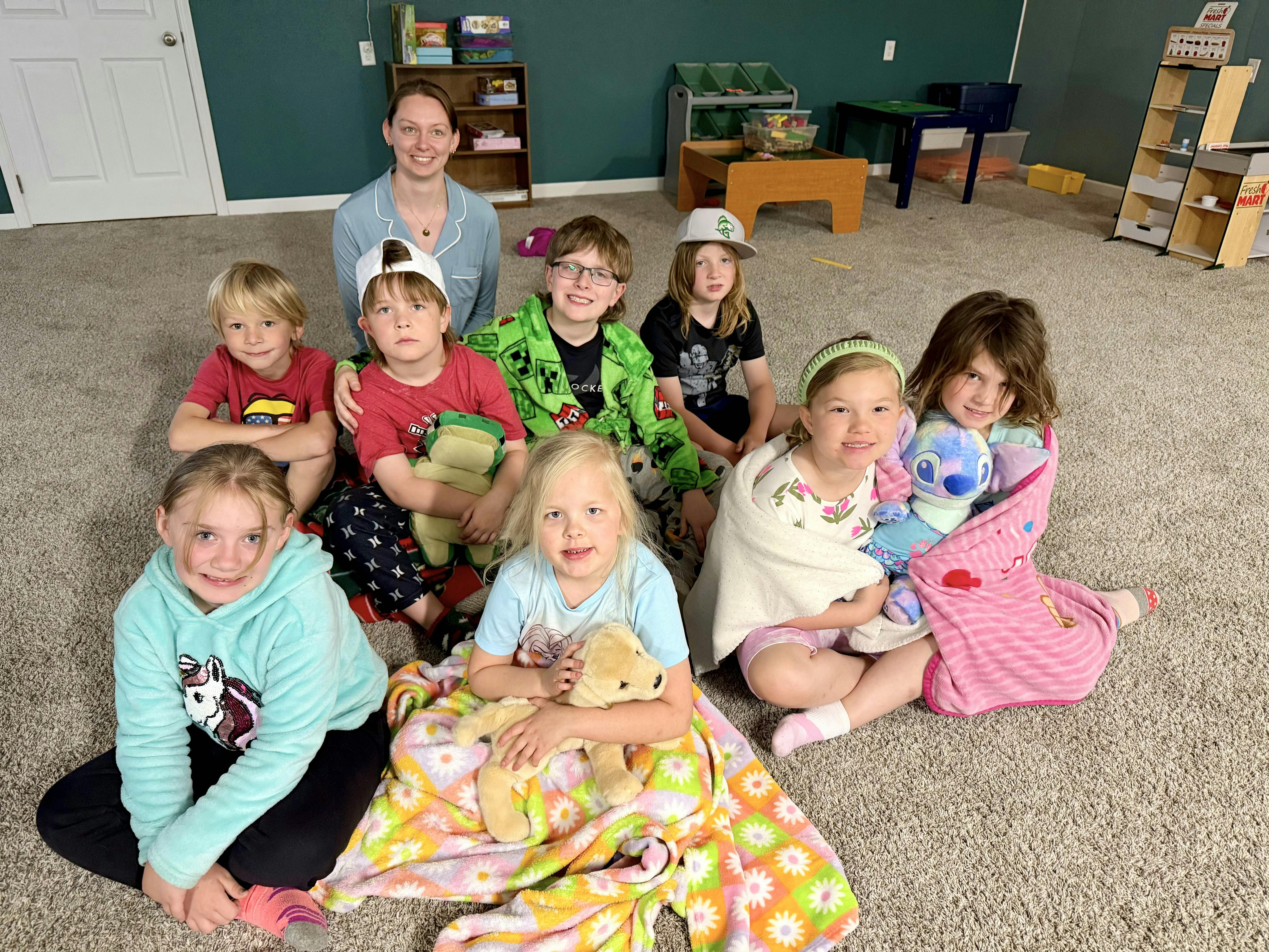 Adult and multiple children sitting on a carpeted floor with blankets and stuffed animals