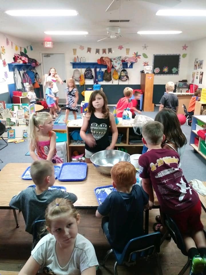 Children in a classroom, some seated at tables, others standing