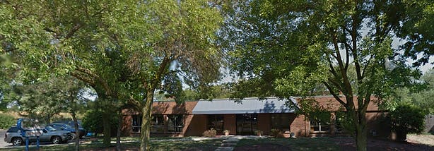 Building with a brown brick facade and a light-colored roof, surrounded by trees