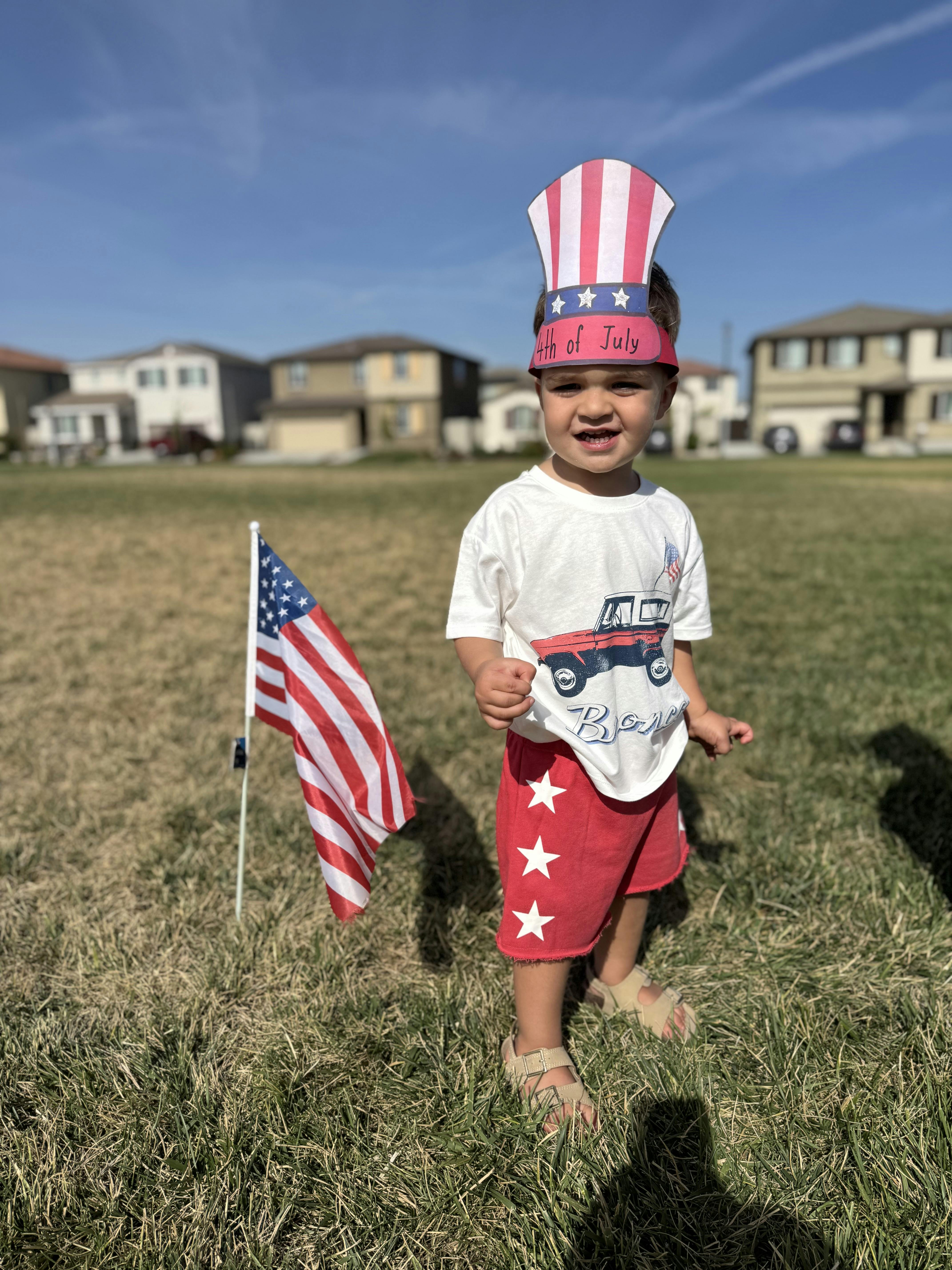 Child wearing a 4th of July hat and shirt standing by an American flag