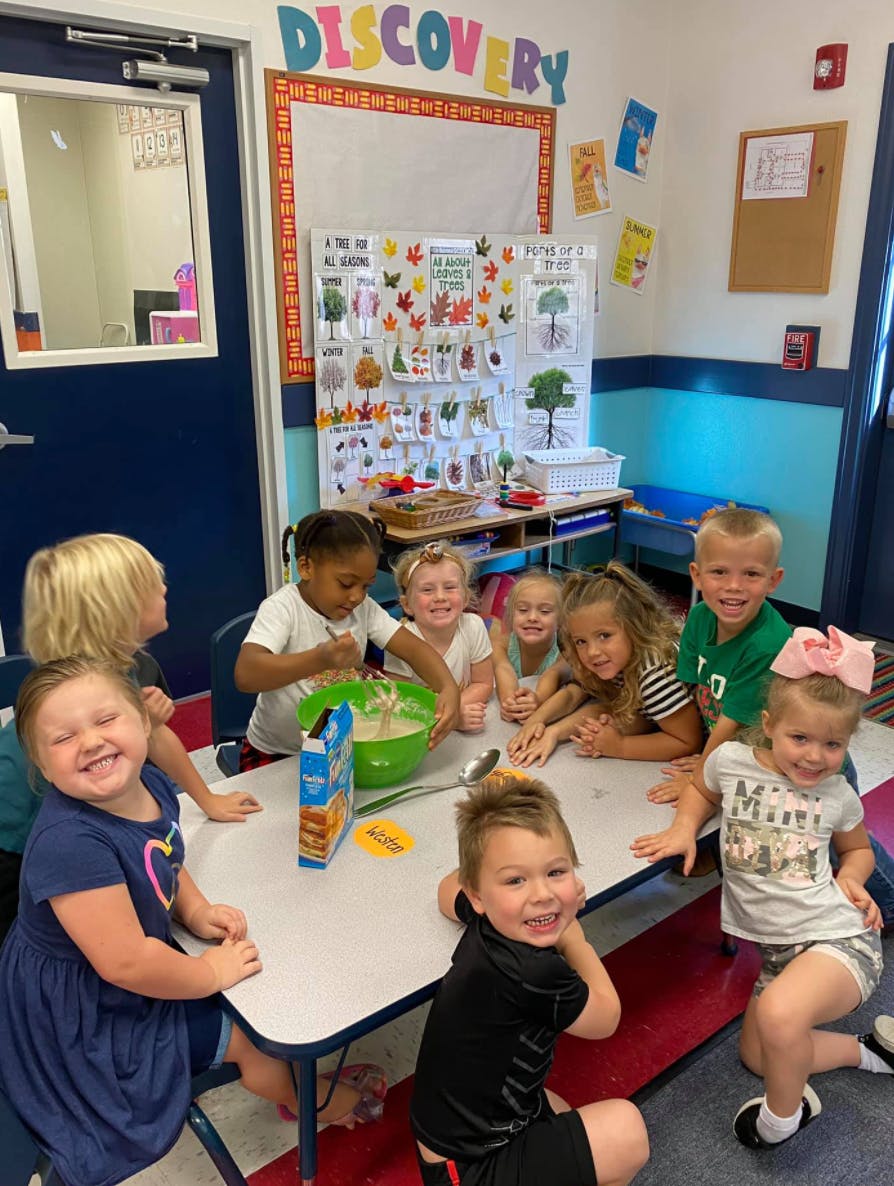 Children gathered around a table in a classroom, one child mixing in a bowl