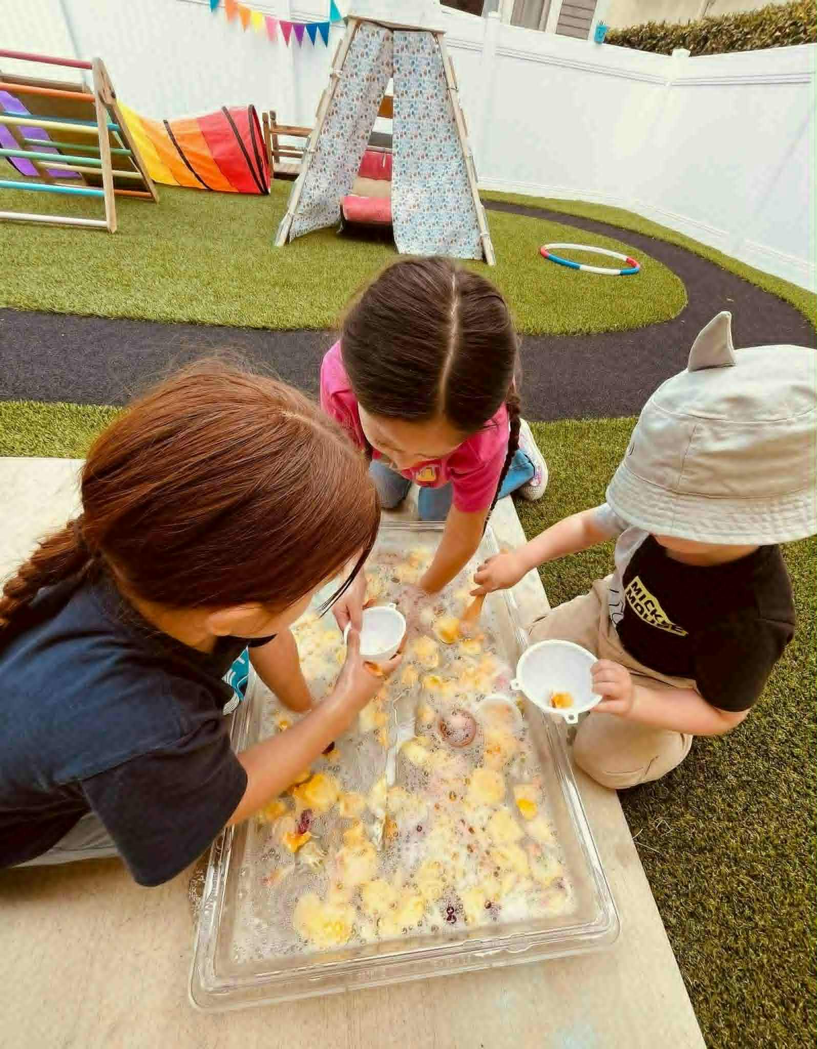 Three children playing with water and fruit in a sensory bin outdoors