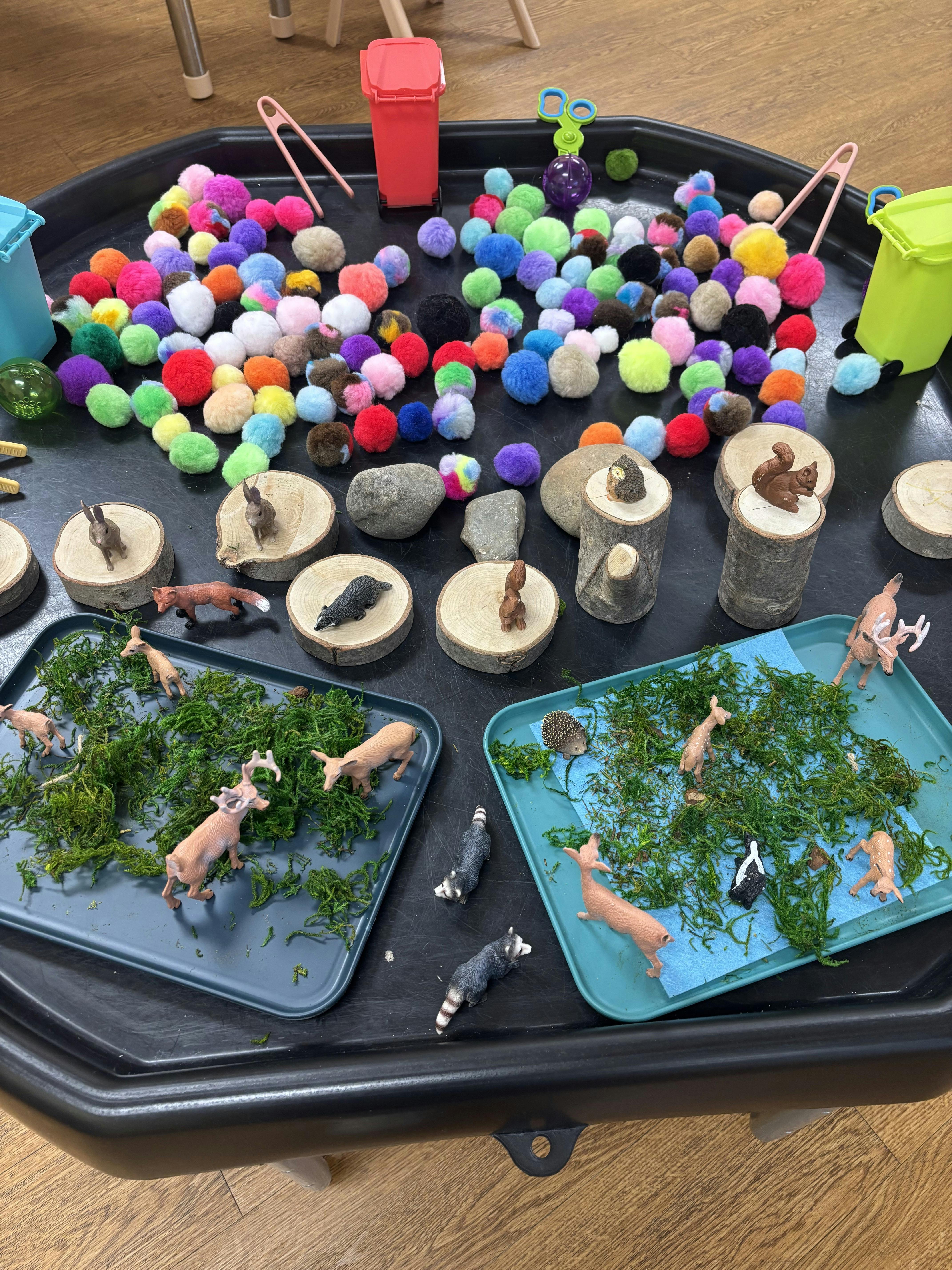 Colorful pom-poms, miniature forest animals, and moss arranged on a black tray
