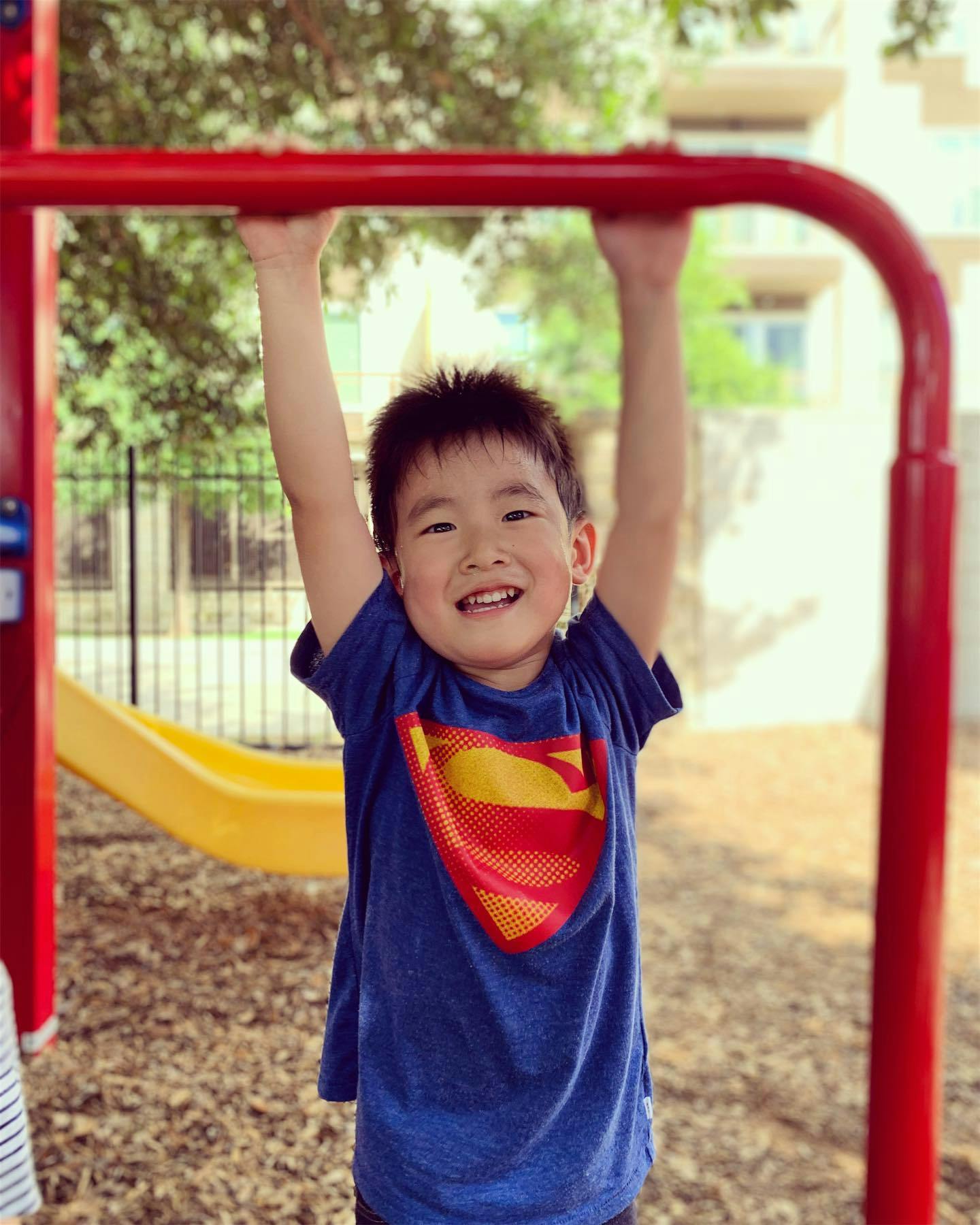 Child in a blue shirt with a Superman logo hanging from red monkey bars