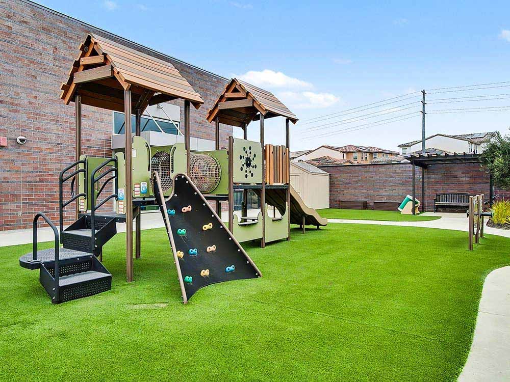 A green and brown playground with slides, a climbing wall, and turf ground