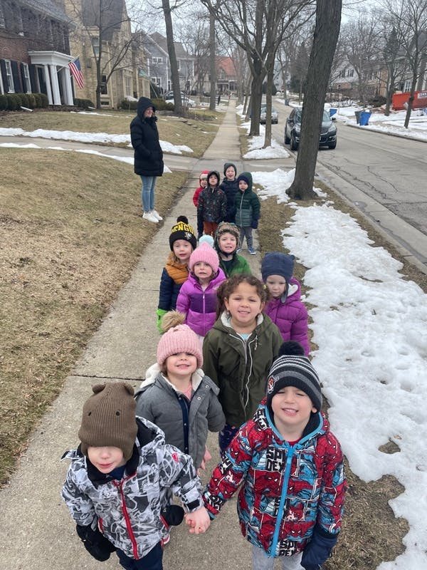 Children walking in a line on a sidewalk with a person observing them