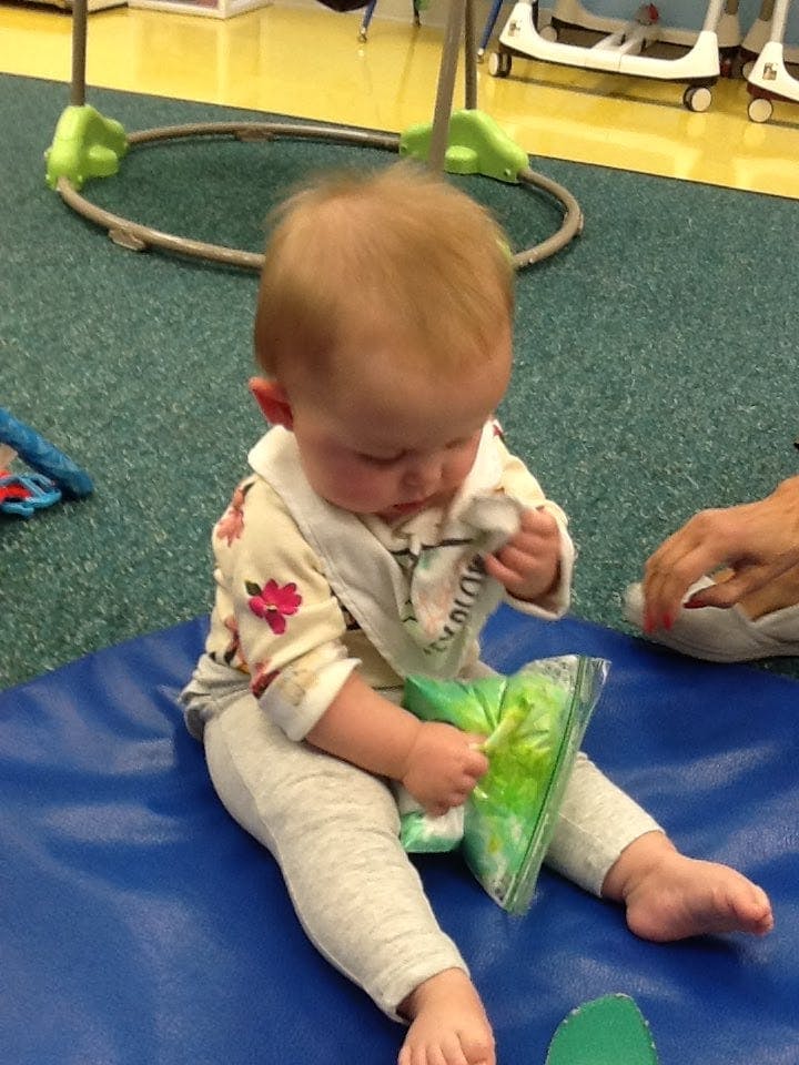 Child sitting on a blue mat, holding a green sensory bag