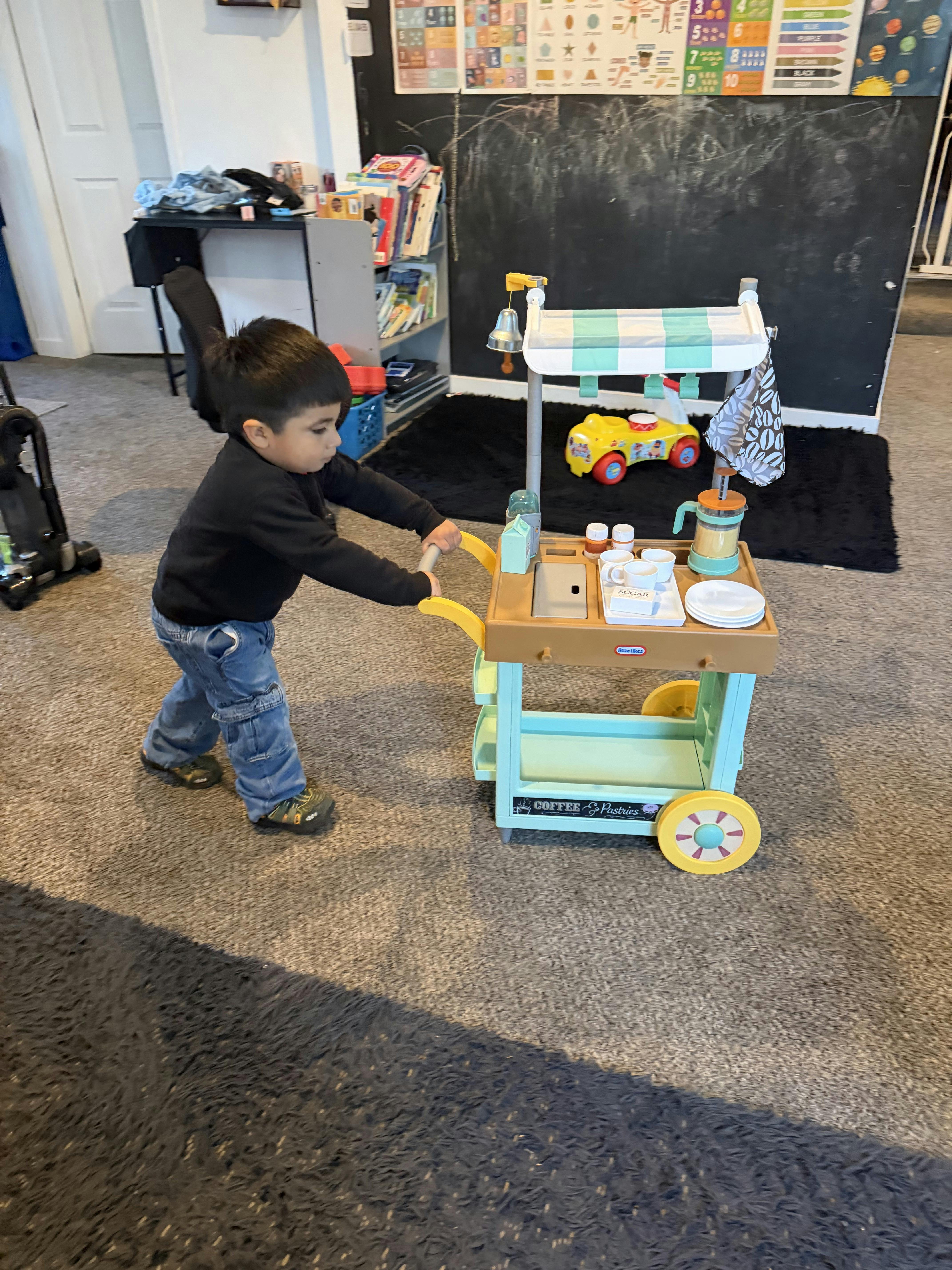 Child pushing a toy coffee cart with cups, plates, and a sign for coffee and pastries