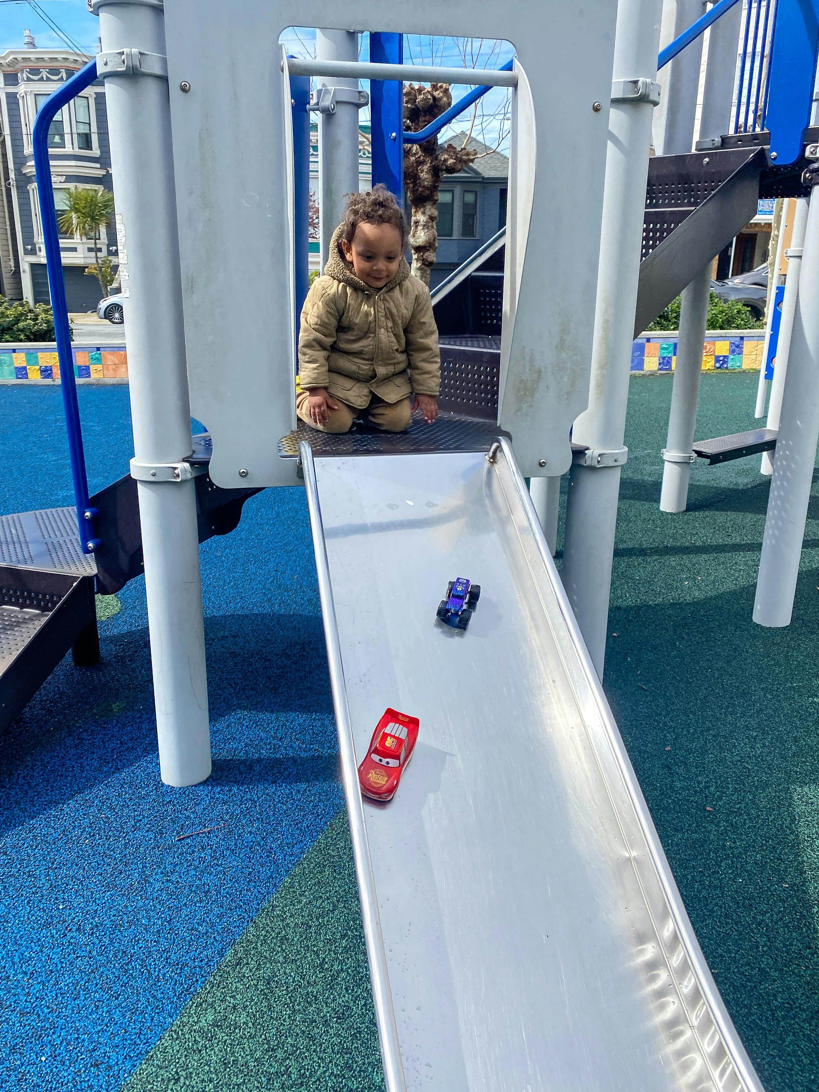 Child kneeling at top of playground slide with toy cars below