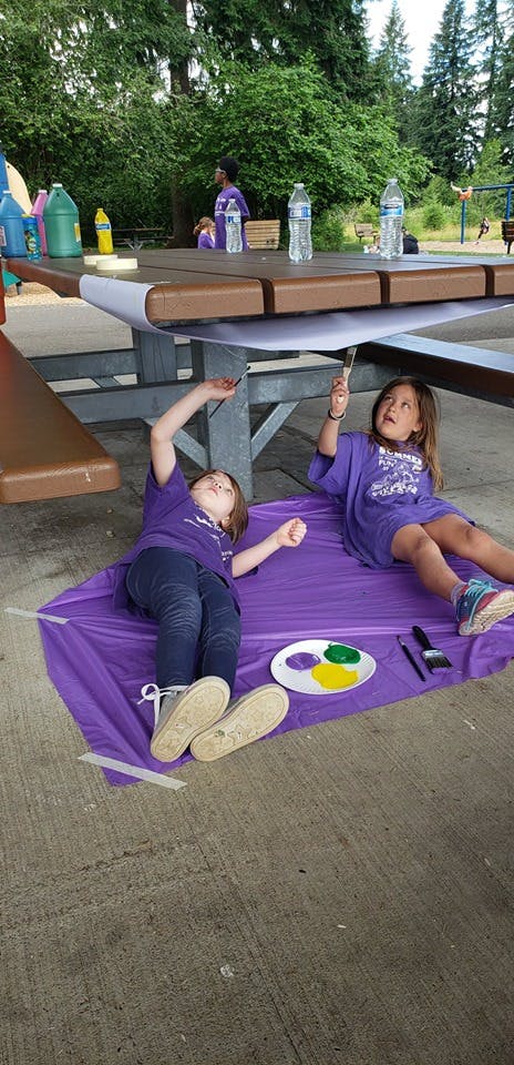 Two children painting on paper under a picnic table
