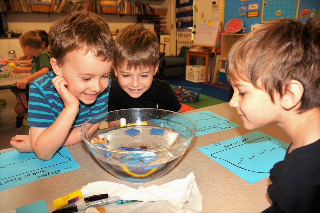 Three children looking at a bowl of water with objects in a classroom