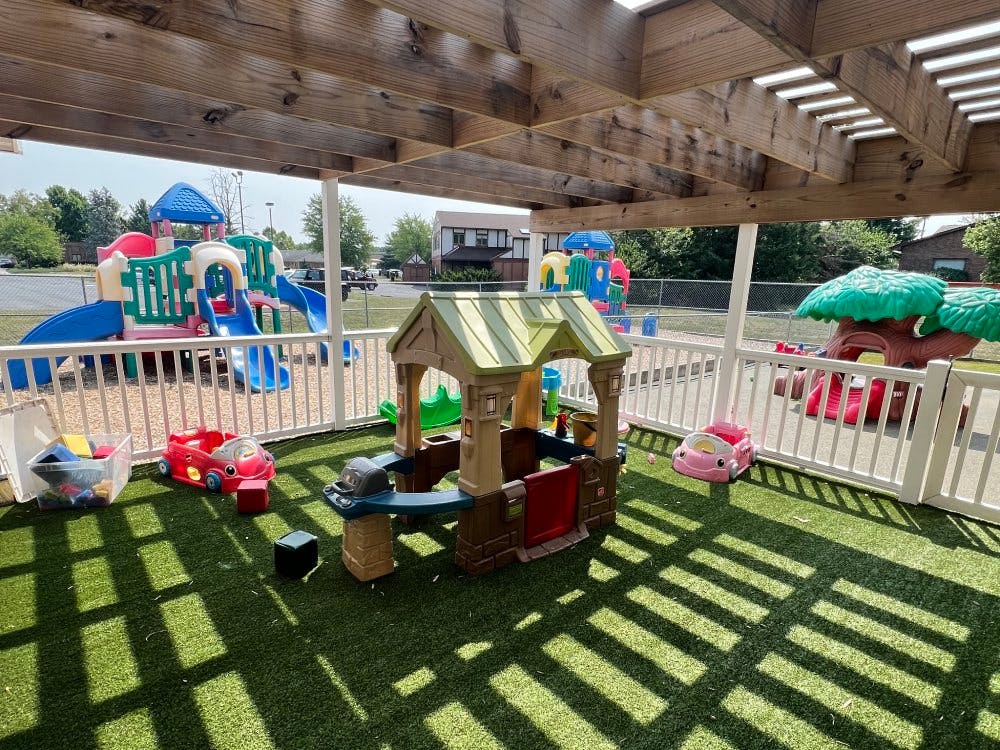 Playground area with playhouses, toy cars, and a play structure beyond a white fence