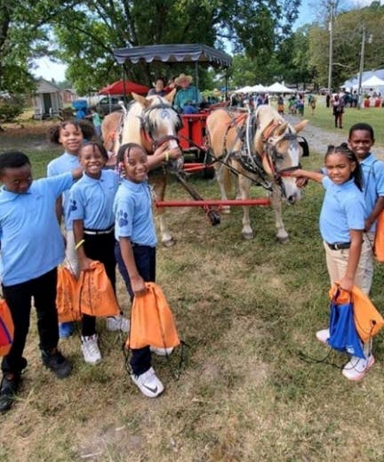 Children in blue shirts holding orange bags next to a horse-drawn carriage