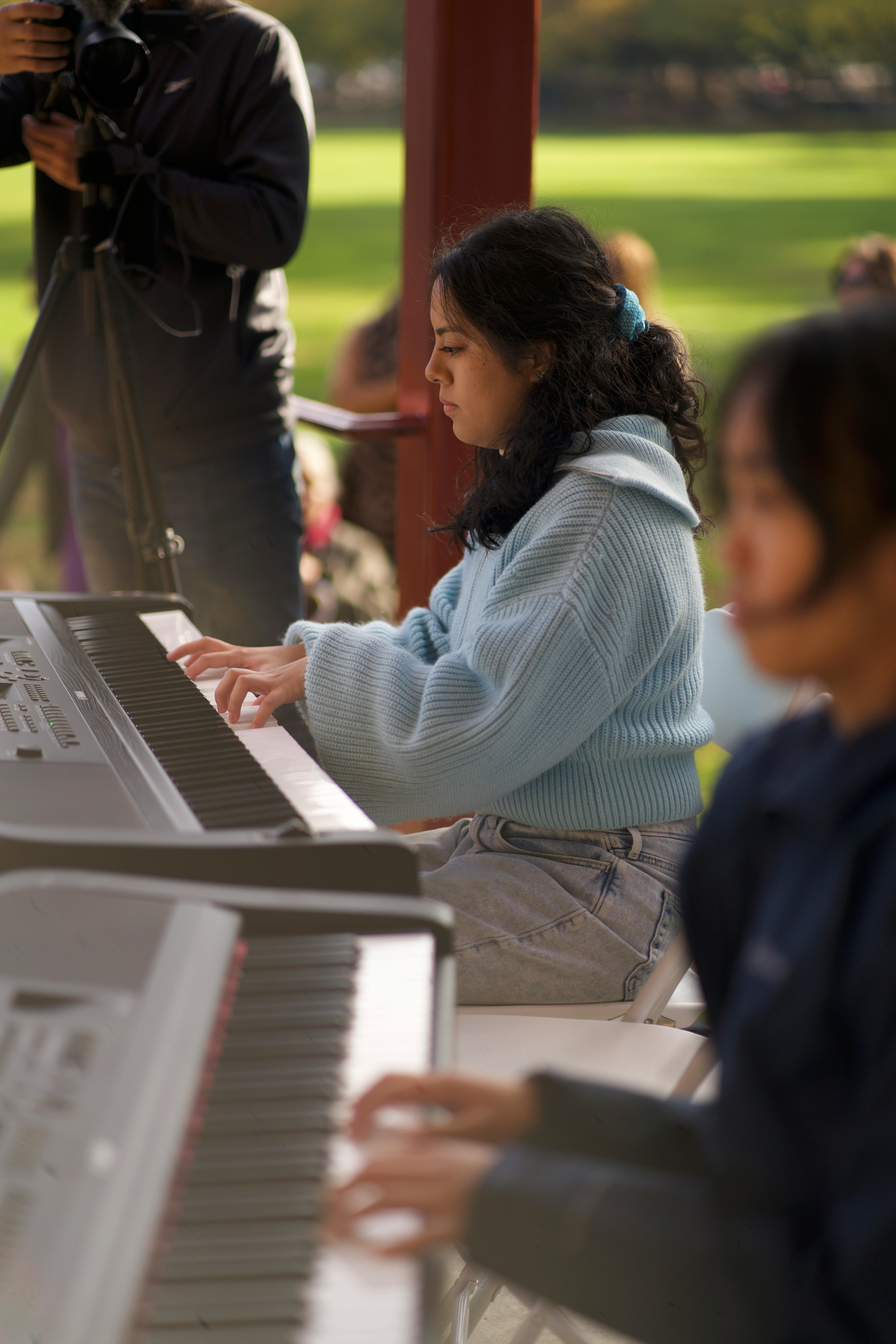 Person playing a keyboard instrument outdoors, with another person in the foreground